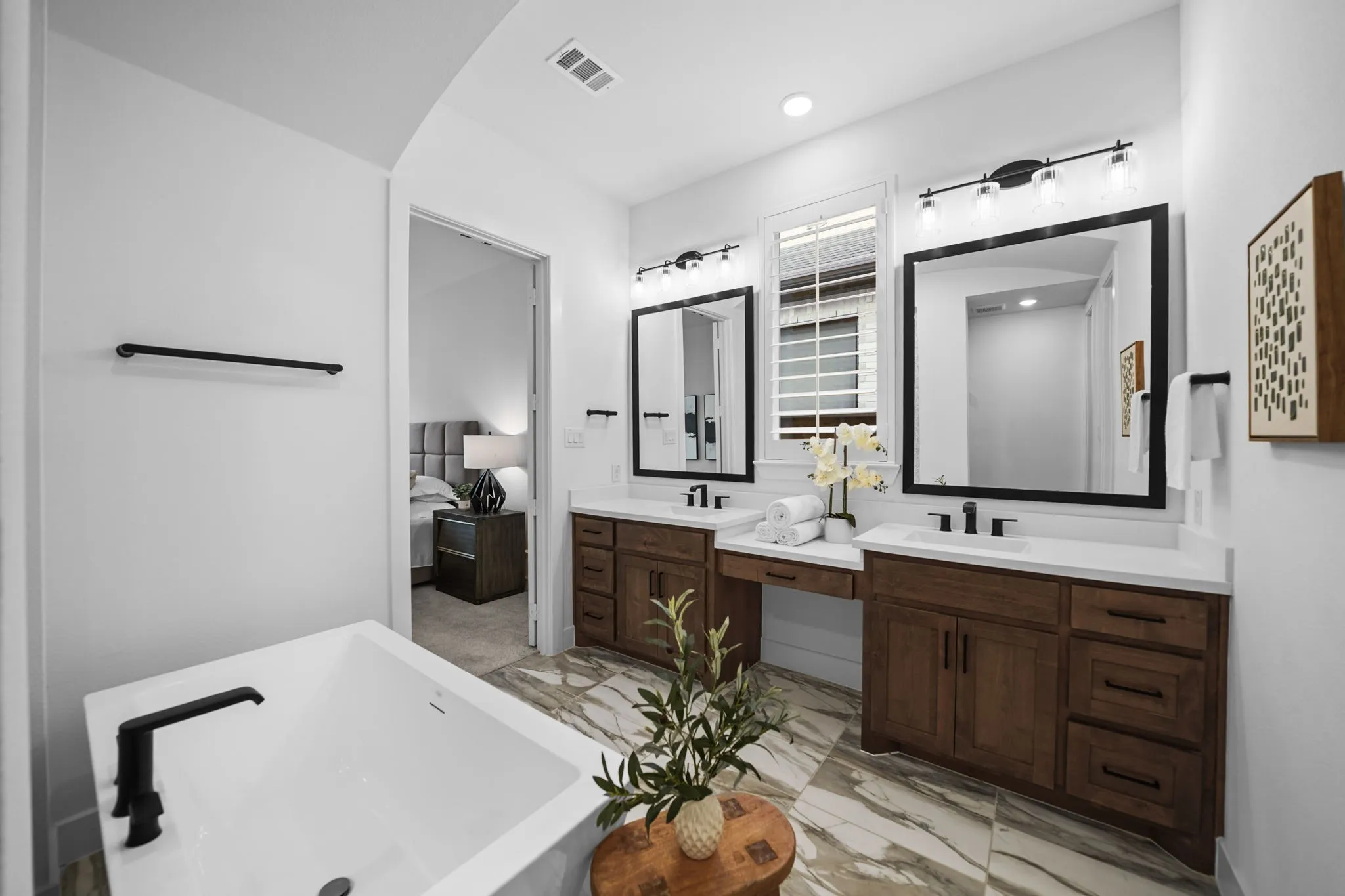 Freestanding soaking tub with black fixtures, dual wood vanities with integrated sinks, framed mirrors with overhead lighting, and patterned floor tiling