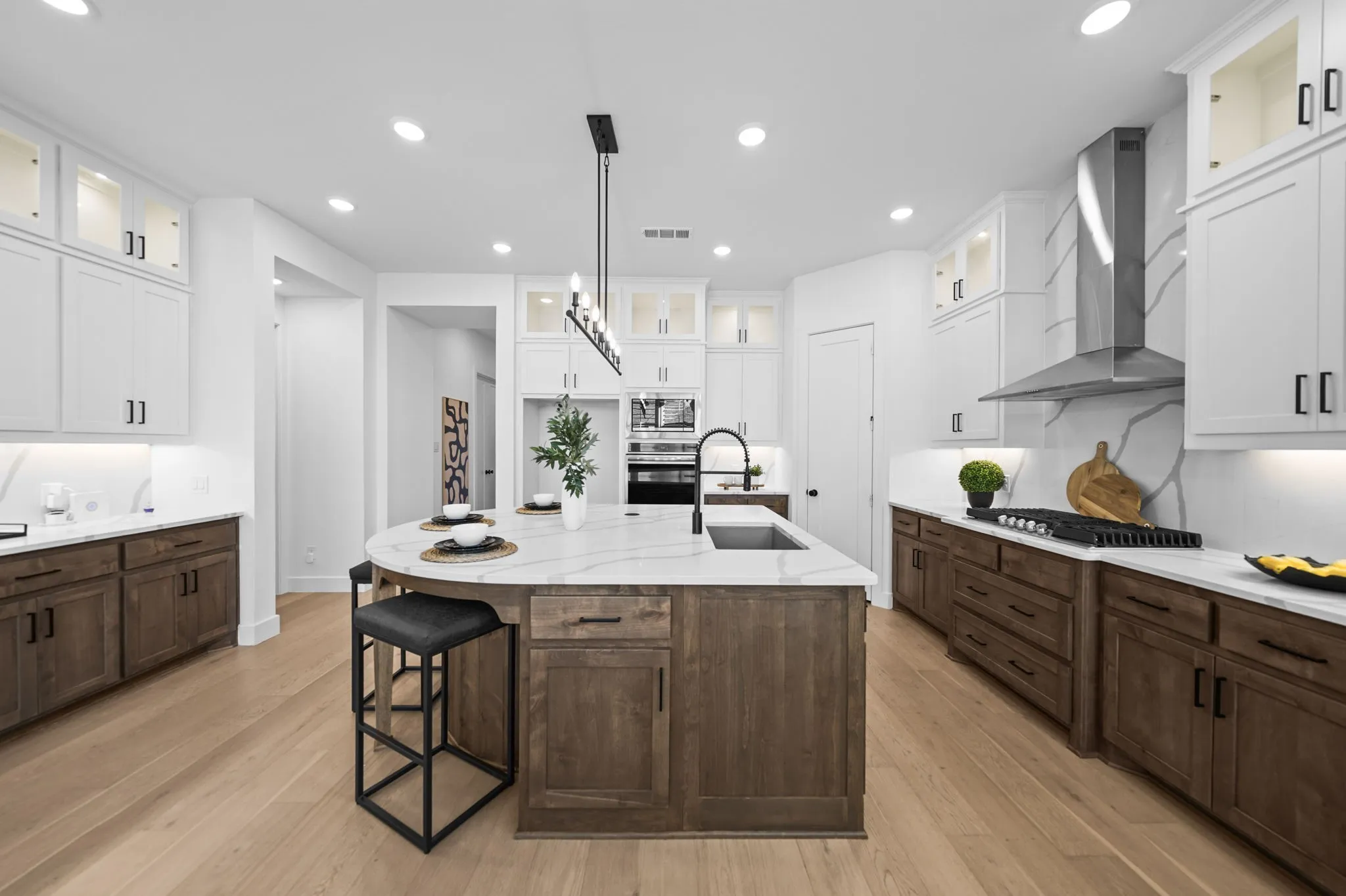 Gourmet kitchen with an expansive island, light wood-finish flooring, white upper cabinetry with glass inserts, dark wood-finish lower cabinetry, and a large stainless steel range hood