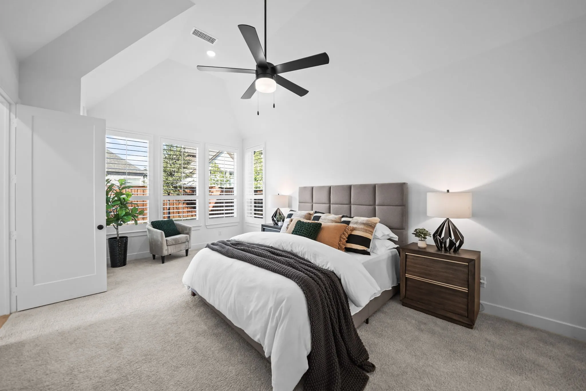 Vaulted ceilings, a dark-toned ceiling fan, multiple windows with plantation shutters, light-toned carpeting, and a flat-panel door with dark hardware