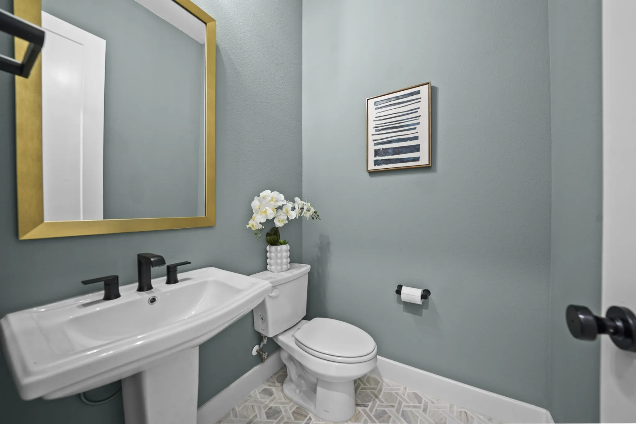 Pedestal sink with a matte black widespread faucet, a gold-framed mirror, and patterned floor tiling