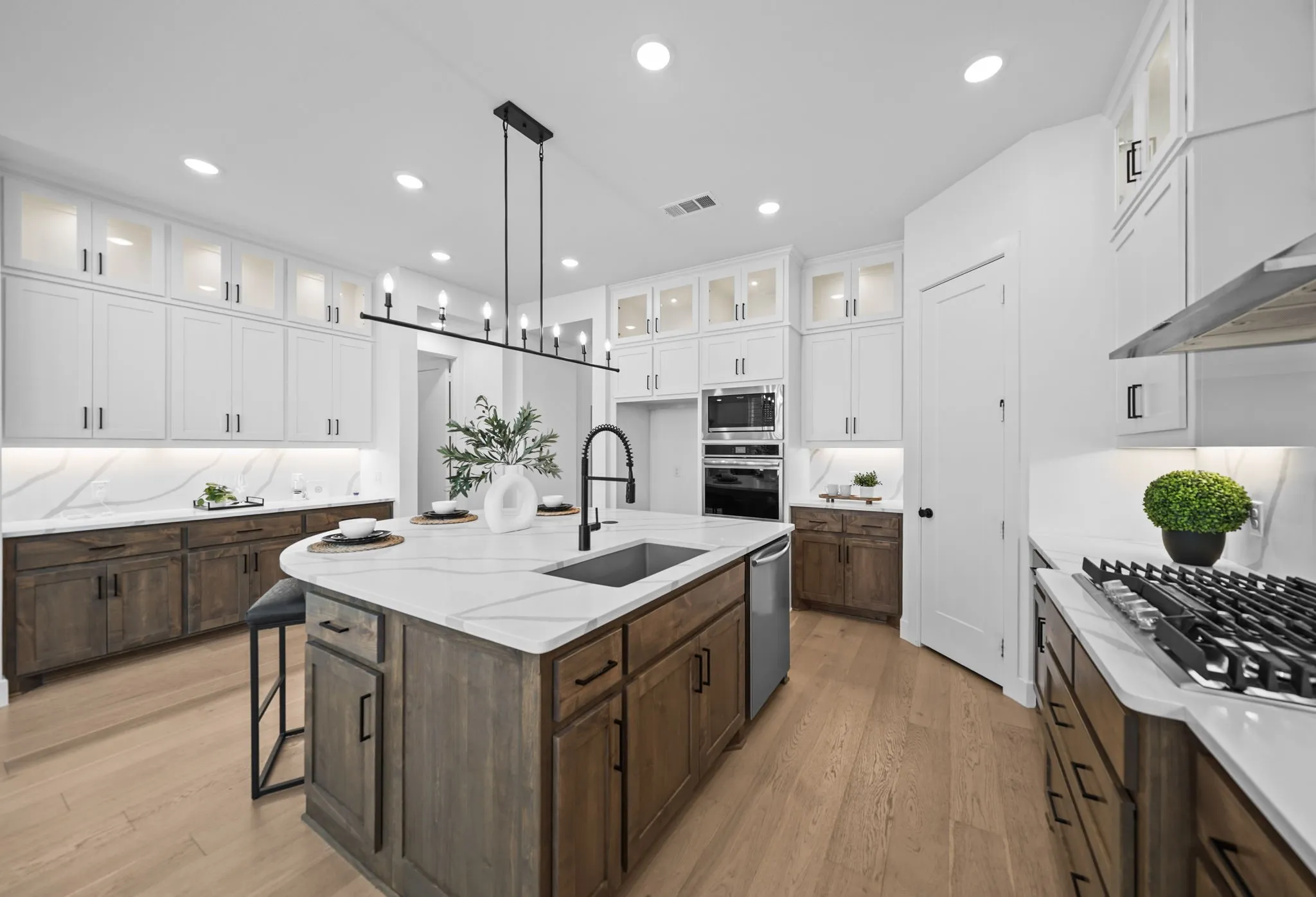 Gourmet kitchen featuring a large central island with an undermount sink, built-in stainless steel appliances, white upper cabinetry with glass inserts, wood-finish lower cabinetry, and light wood-finish flooring