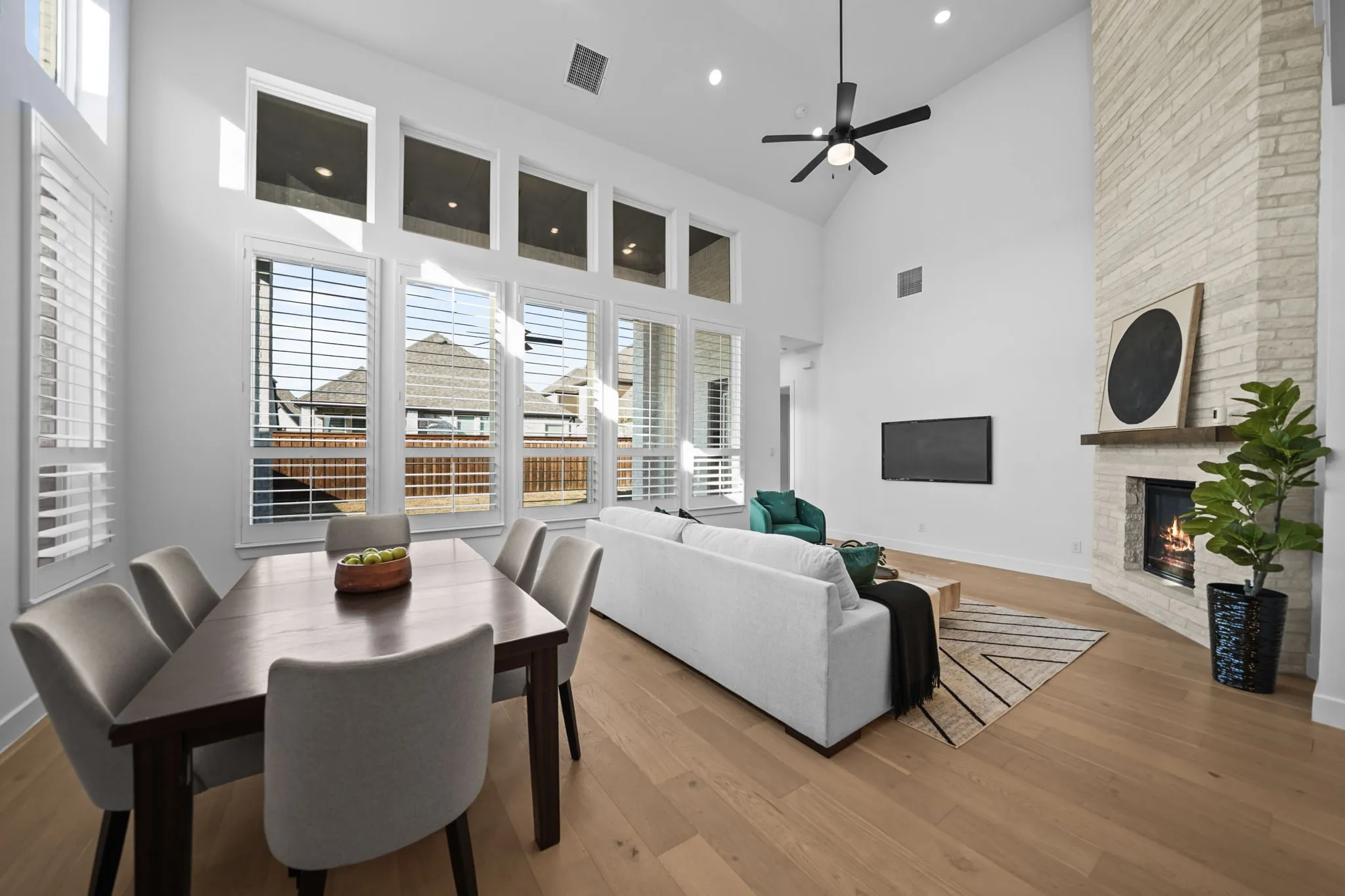 Vaulted ceilings, a stone-clad fireplace with a wood mantel, wood-finish flooring, a ceiling fan, and extensive windows with plantation shutters