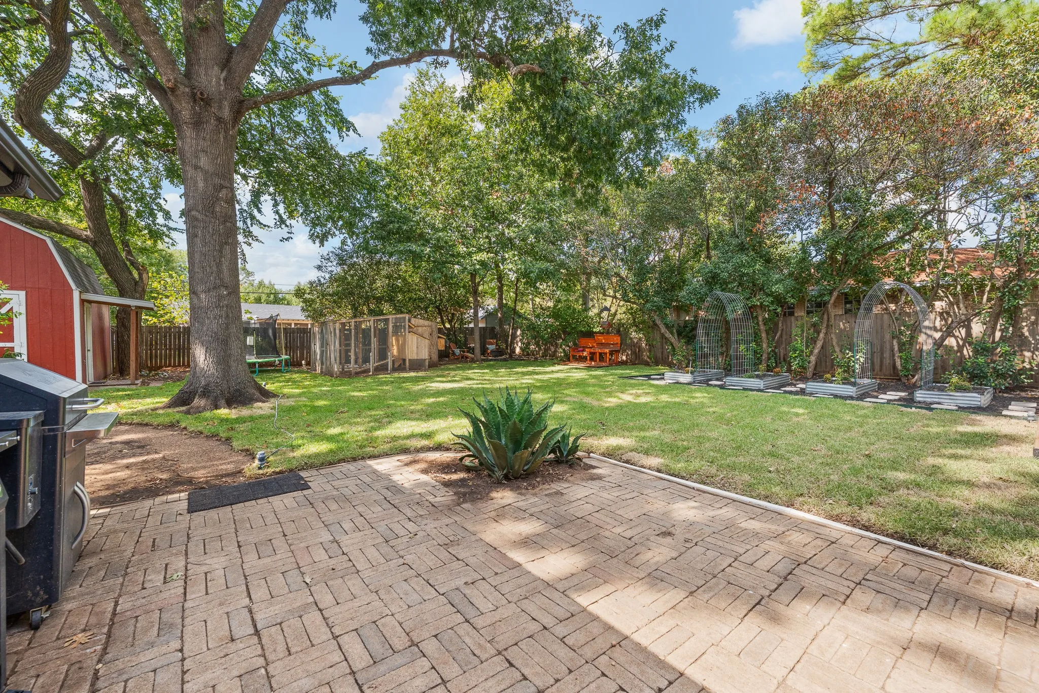 Spacious backyard featuring a brick paver patio, a large mature tree, a red storage shed, and a fenced garden area