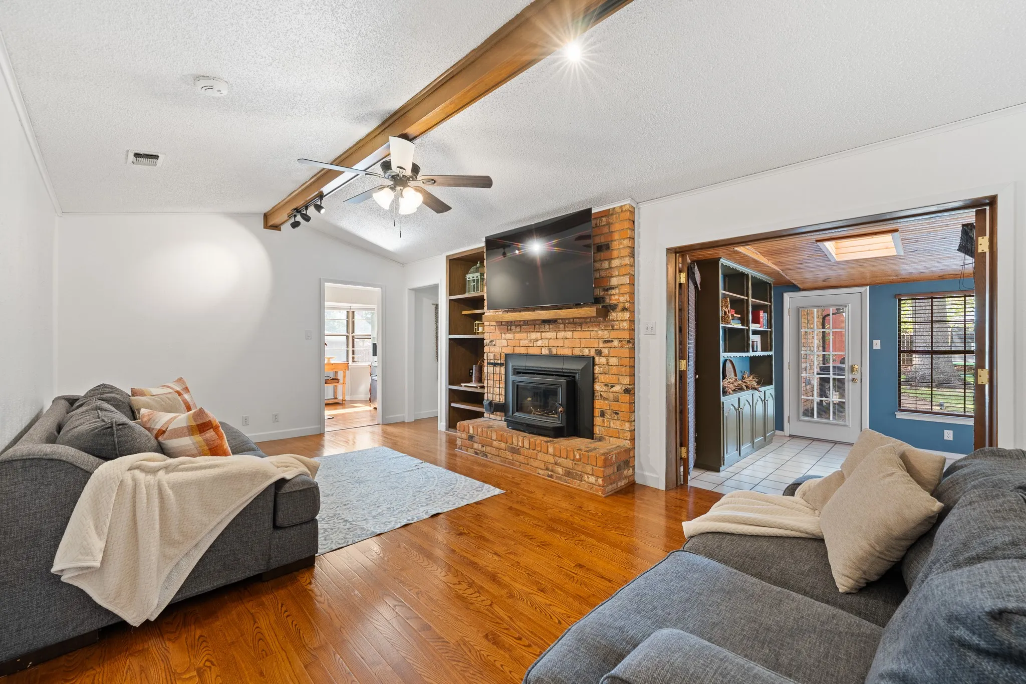 Vaulted ceiling with wood beam accent, brick fireplace with built-in shelving, wood-finish flooring, and ceiling fan