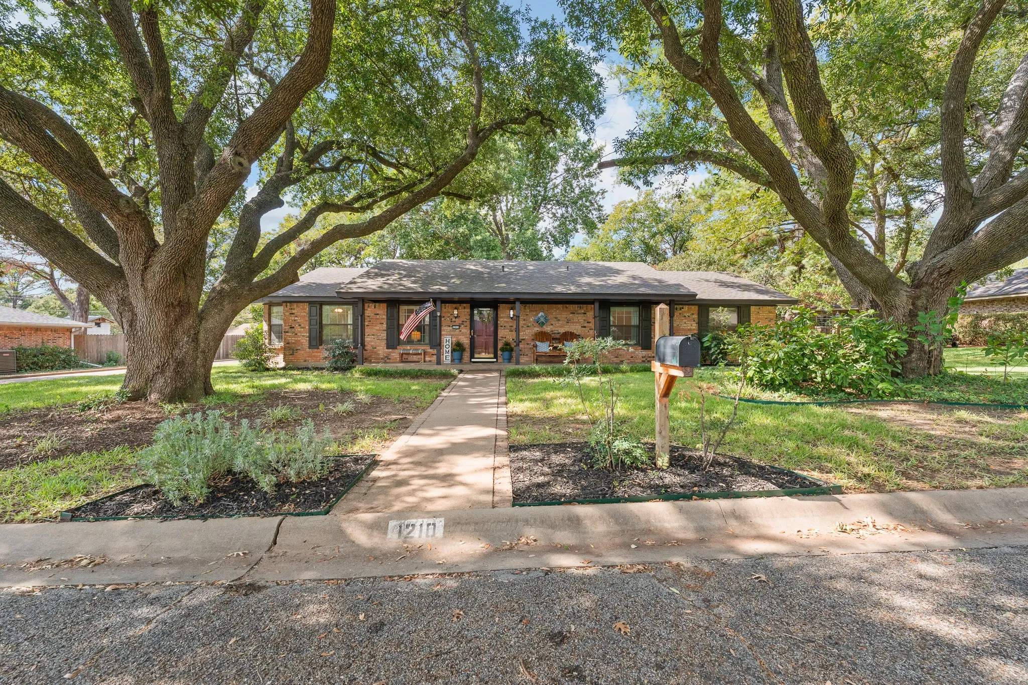 Brick exterior ranch home with a shingled roof, mature trees, a concrete walkway, and landscaped garden beds
