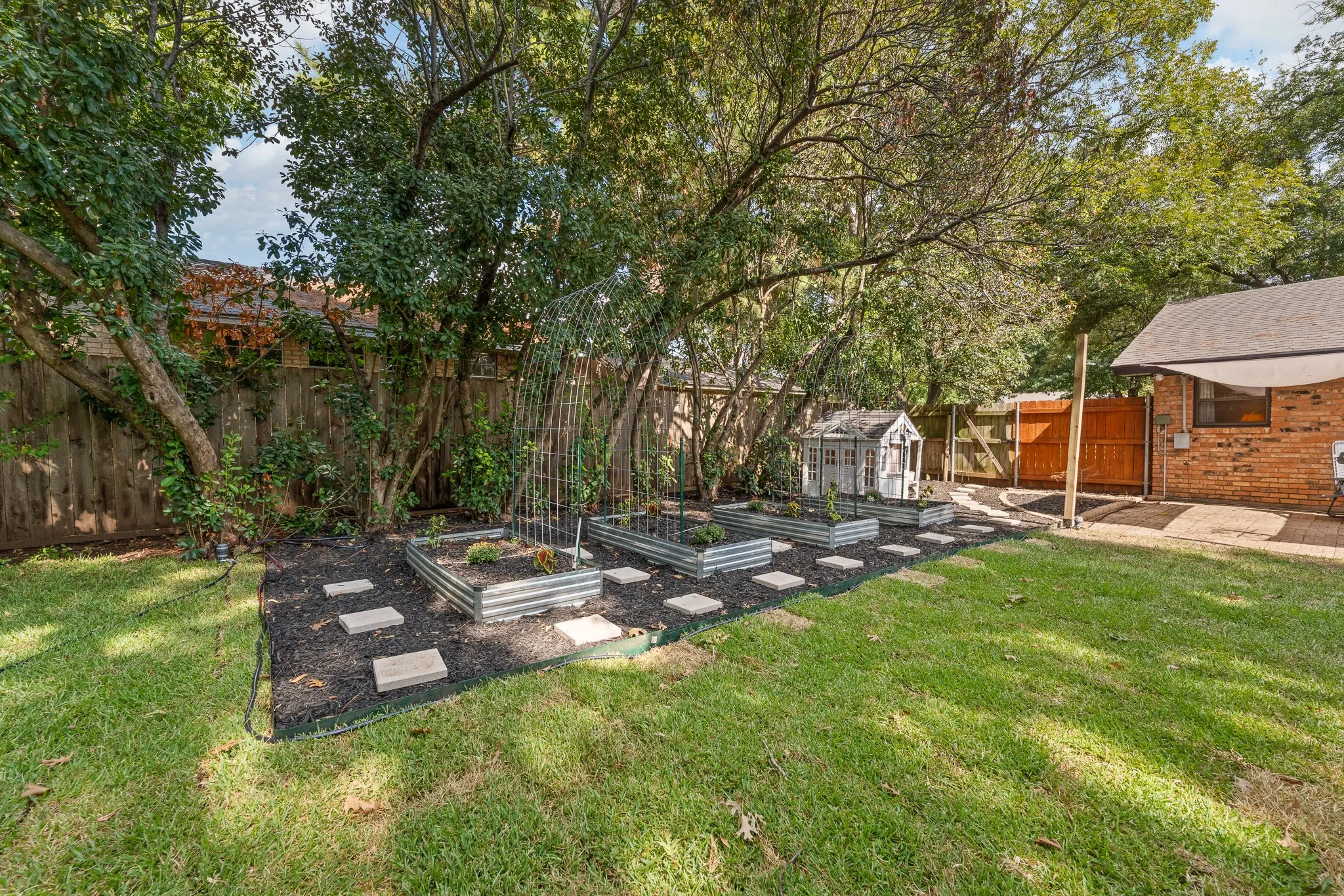 Manicured lawn with mature trees, raised garden beds, stepping stones, and a brick exterior structure