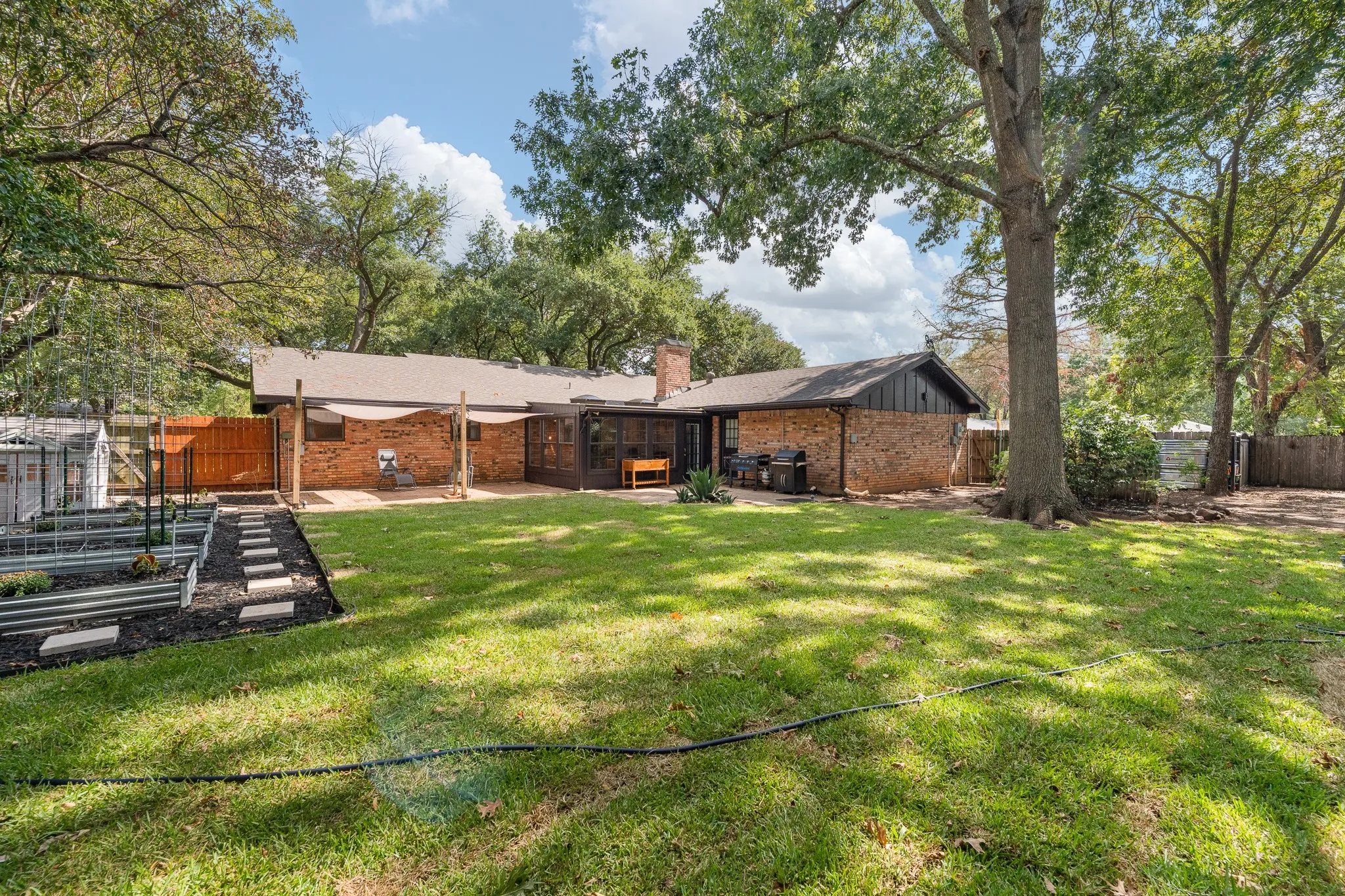 Expansive backyard featuring a manicured lawn, established shade trees, and a brick exterior home with a covered patio area