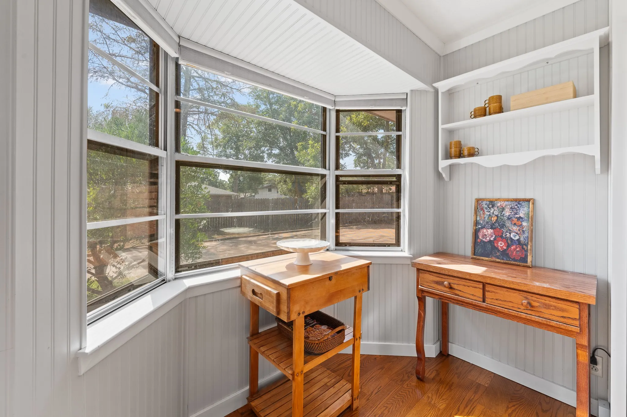 Bay window with clear glass panels, wood-finish flooring, white beadboard wall paneling, and white built-in shelving