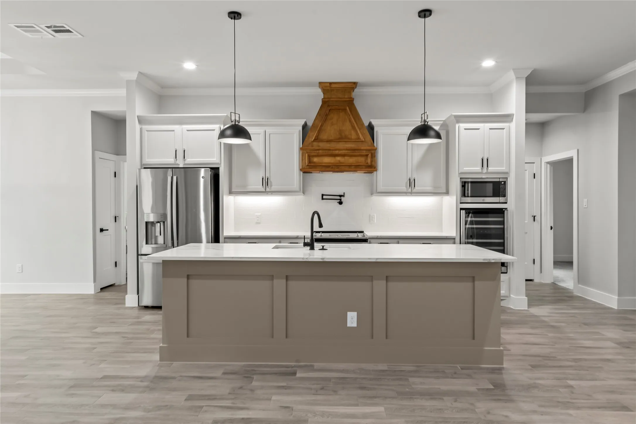 Spacious kitchen featuring a central island with a light-toned base and a white countertop, stainless steel appliances, a decorative wood range hood, and pendant lighting