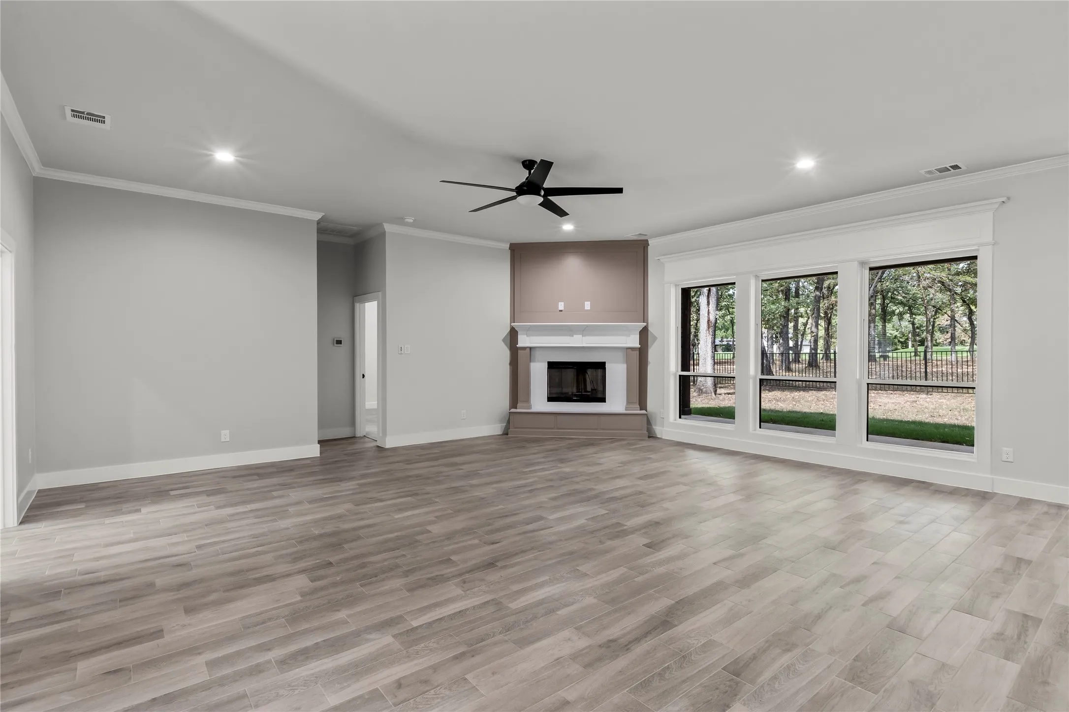 Spacious room featuring wood-finish flooring, a corner fireplace with white mantle, crown molding, a ceiling fan, and large windows with exterior landscaping