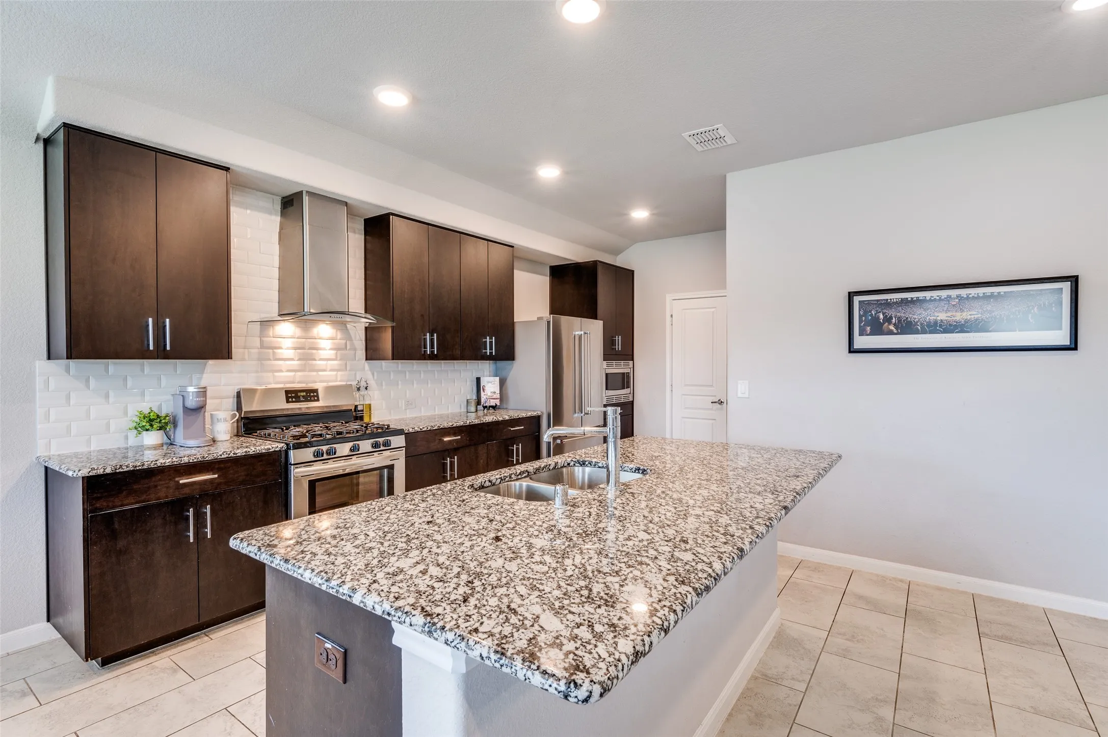 Kitchen featuring dark wood-finish cabinetry, stainless steel appliances, a white subway tile backsplash, light-toned tile flooring, and a large central island with a granite countertop and integrated sink