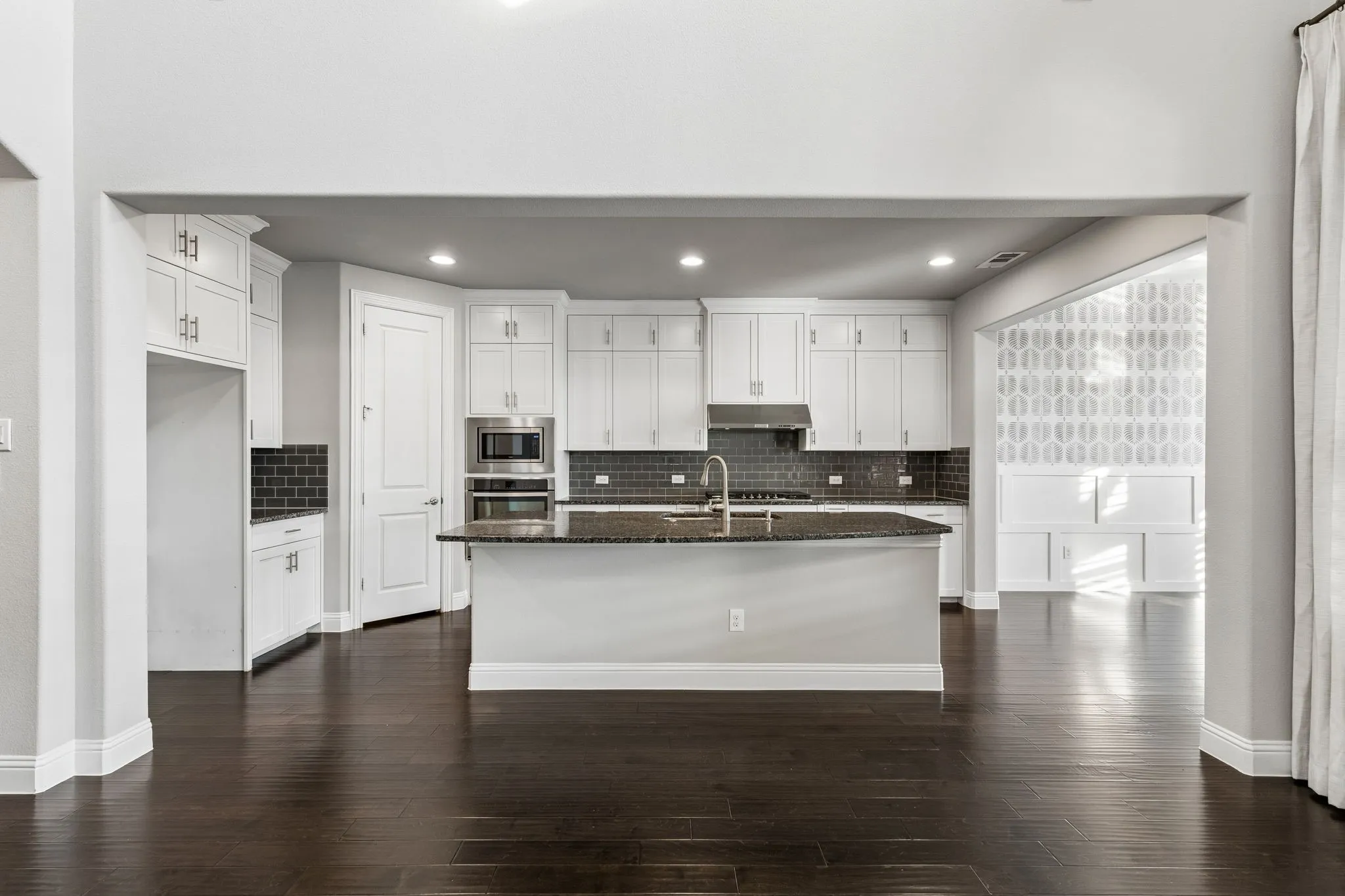 The open-concept kitchen is anchored by an expansive granite island and surrounded by ceiling-height white shaker cabinetry, stainless appliances and rich hardwood floors