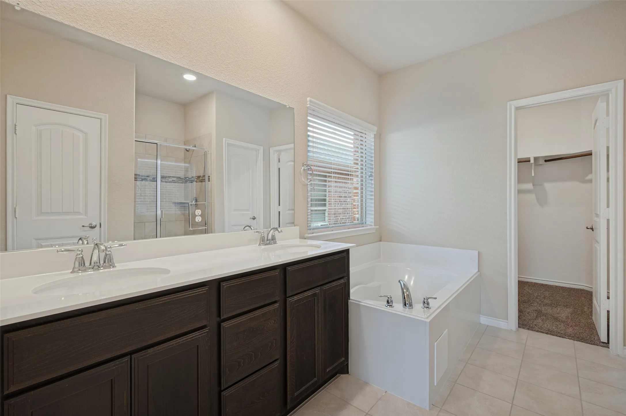 Dual vanity with undermount sinks and chrome faucets, large mirror, built-in bathtub with chrome fixtures, tiled flooring, and a walk-in closet with shelving