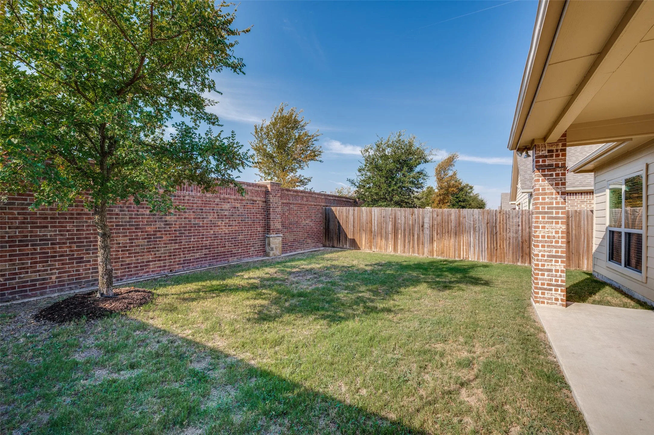 Expansive backyard featuring a manicured lawn, brick perimeter wall, wood privacy fence, and a covered patio area