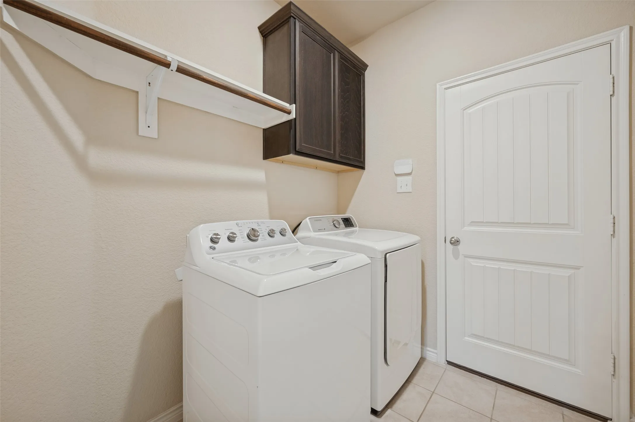 Laundry room featuring a dark wood-finish cabinet, a white shelf with a hanging rod, and light-toned tile flooring