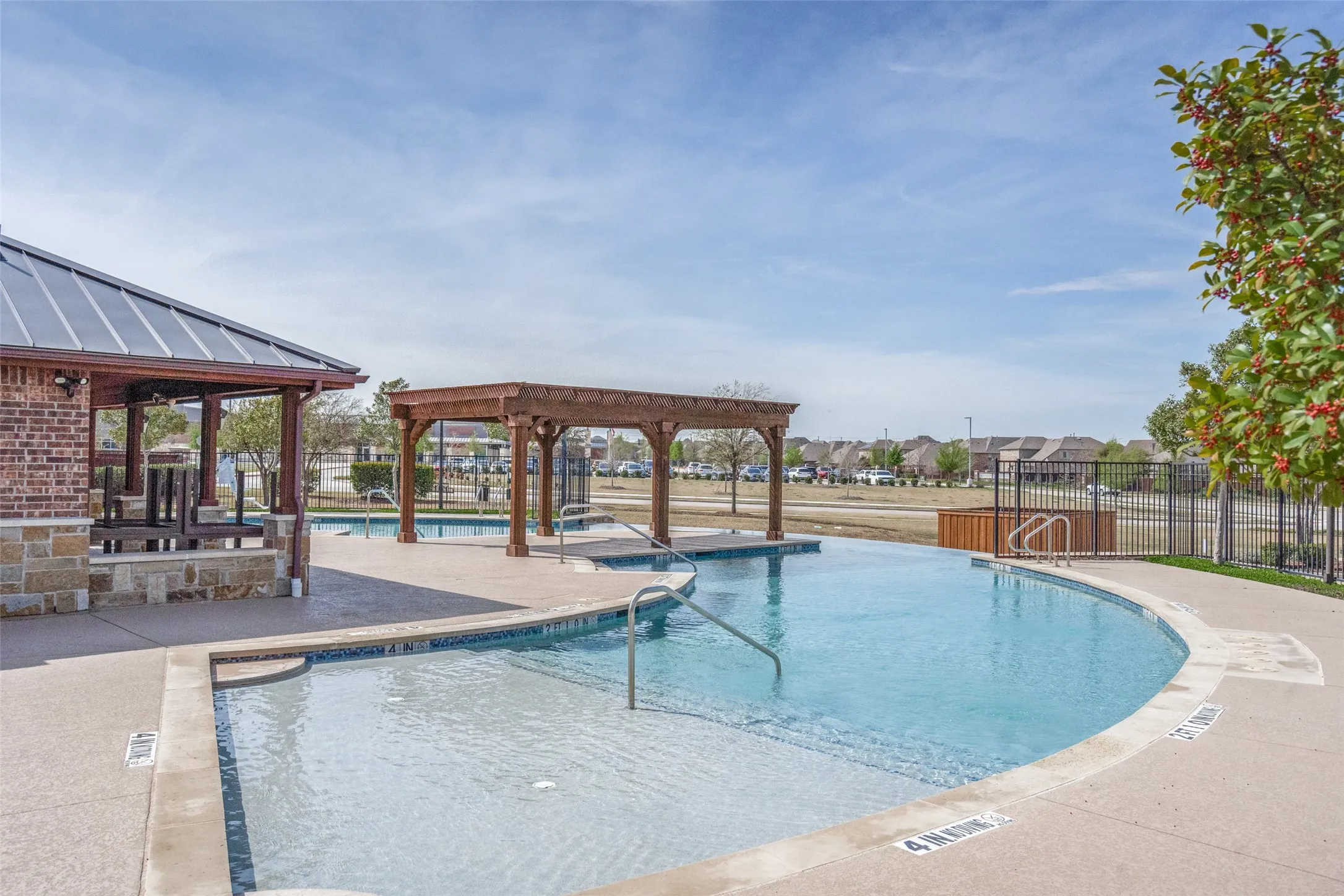 Curved swimming pool with a shallow entry, adjacent brick pavilion with a metal roof, and a wooden pergola structure