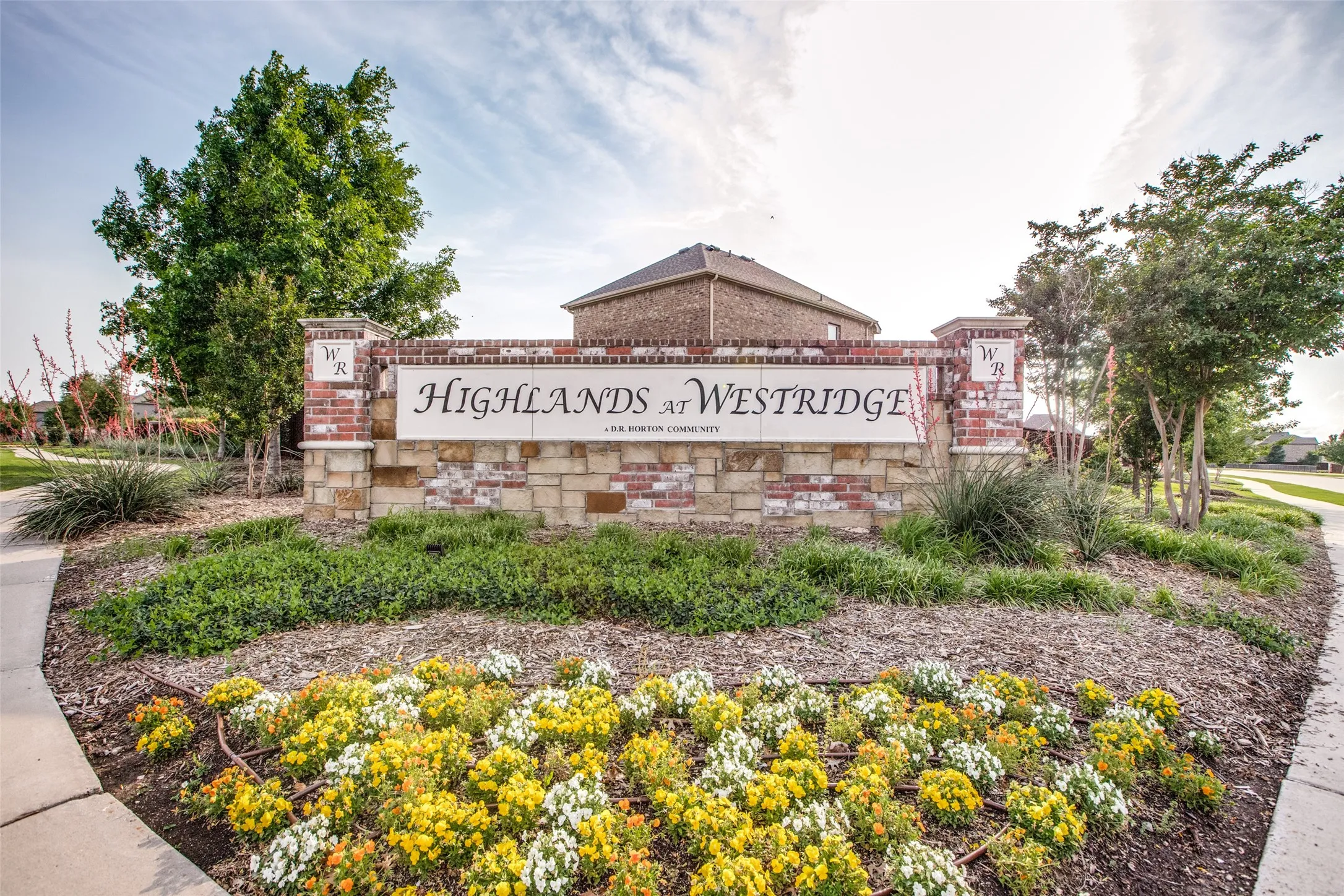 Stone and brick monument sign with raised lettering, surrounded by varied landscaping, mature trees, and a concrete pathway