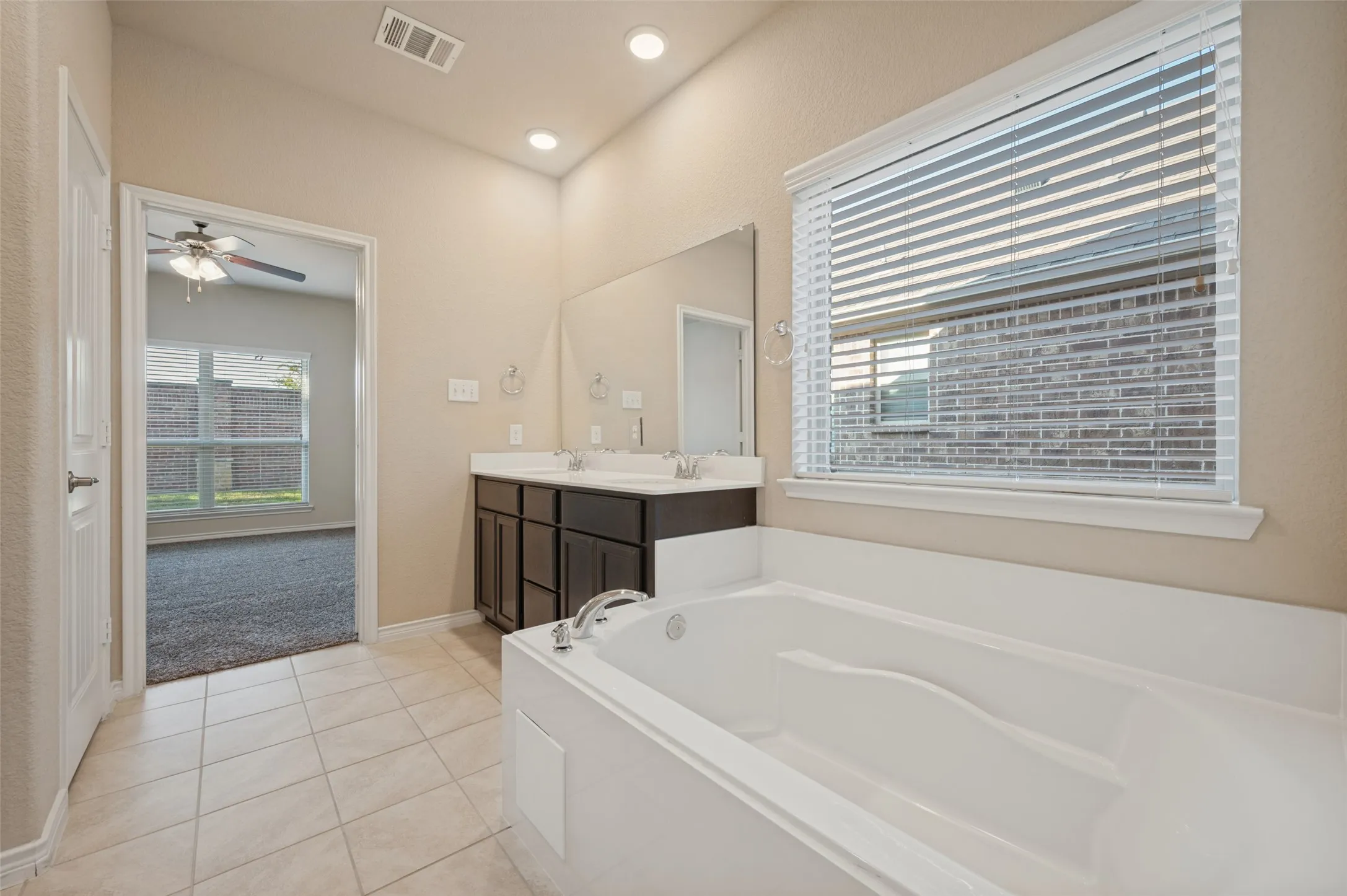 Soaking tub, dual vanity with undermount sinks, large wall-mounted mirror, ceramic tile flooring, and a window with blinds