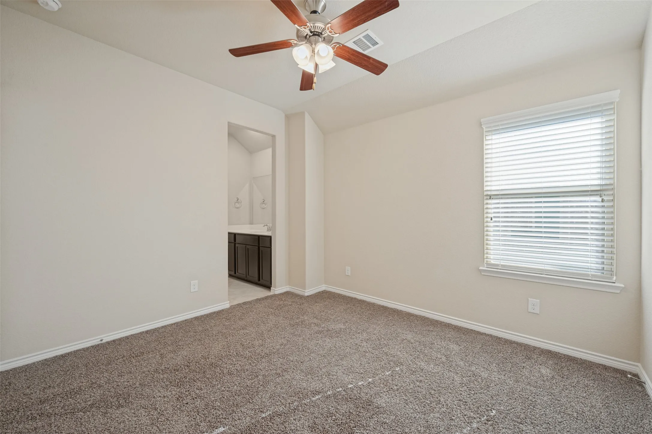 Carpeted room with a ceiling fan light fixture, window with blinds, and an attached bathroom featuring a dark wood-finish vanity