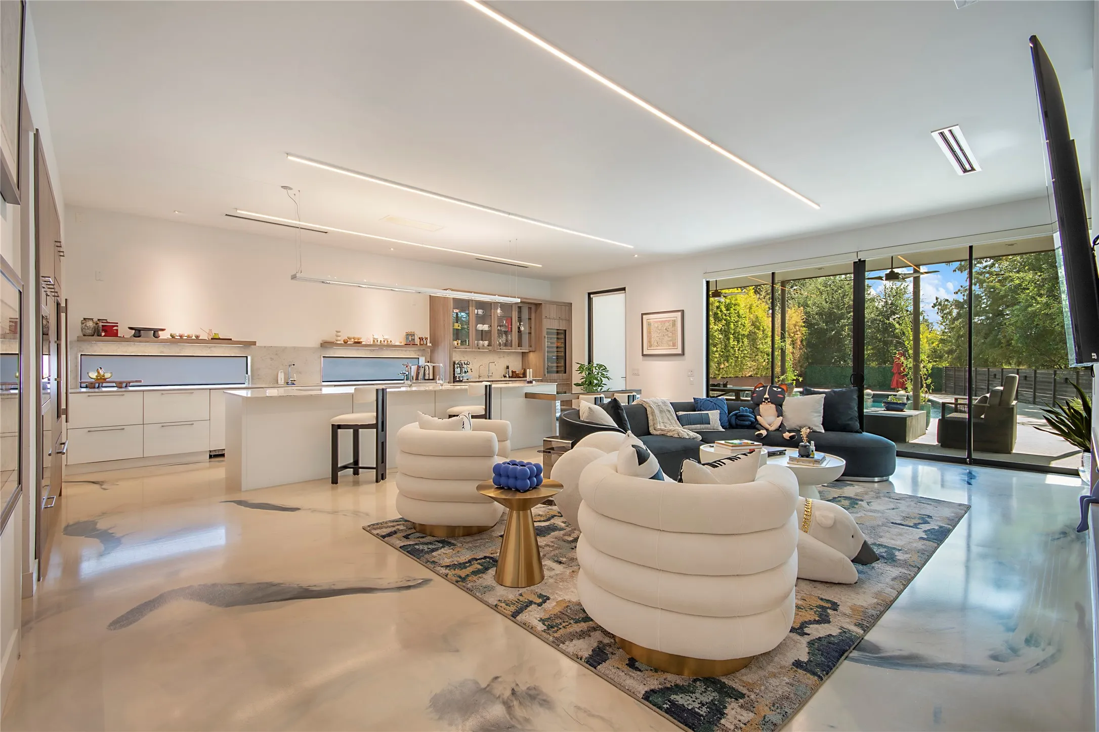 Contemporary open-concept living space featuring polished concrete flooring, seamless floor-to-ceiling glass walls with natural light, integrated linear ceiling lighting, custom built-in cabinetry with display shelving, and direct access to landscaped exterior grounds