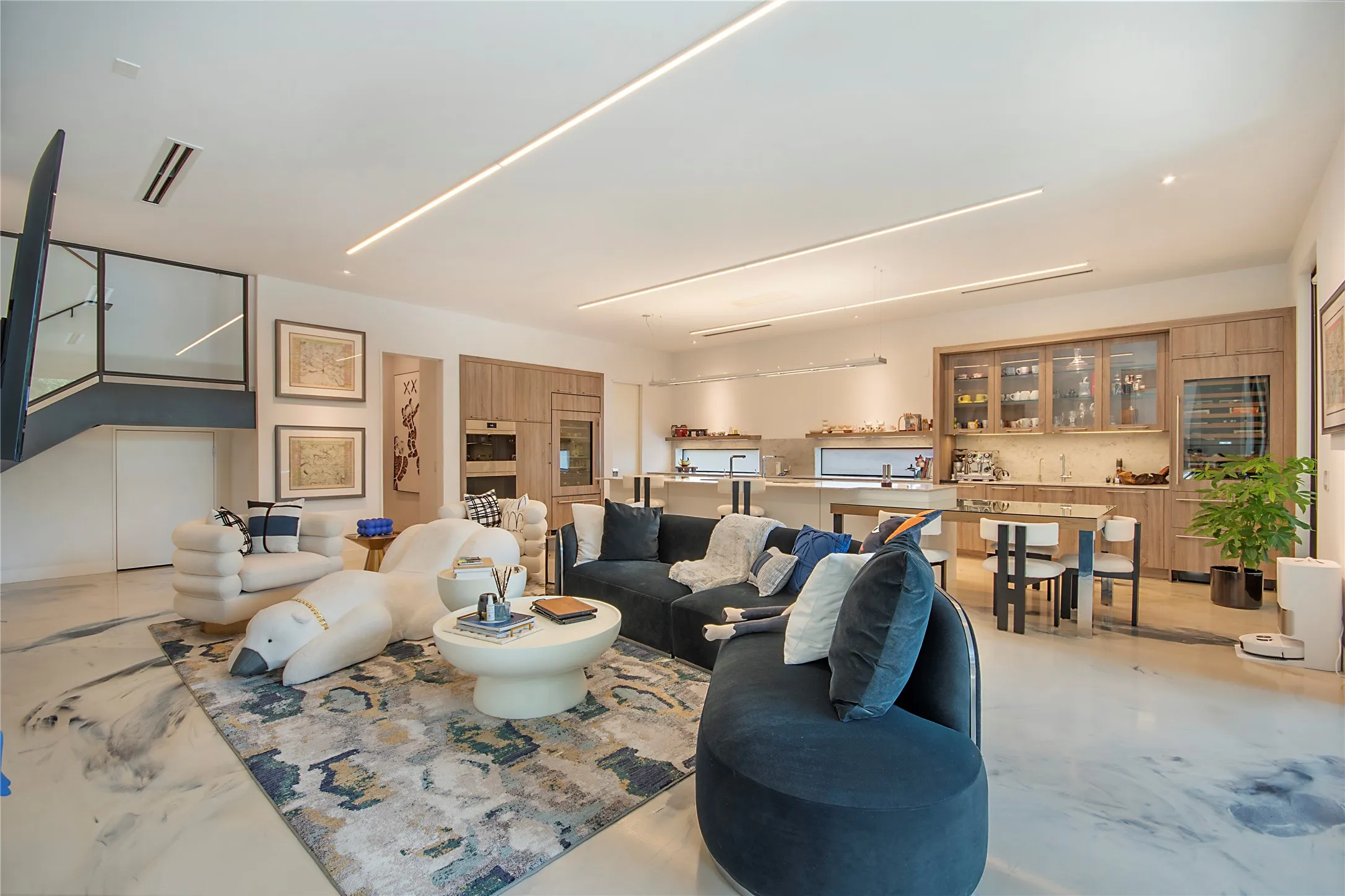 Open-concept living space with sleek linear lighting, built-in kitchen cabinetry, integrated appliances, and a contemporary glass and metal staircase