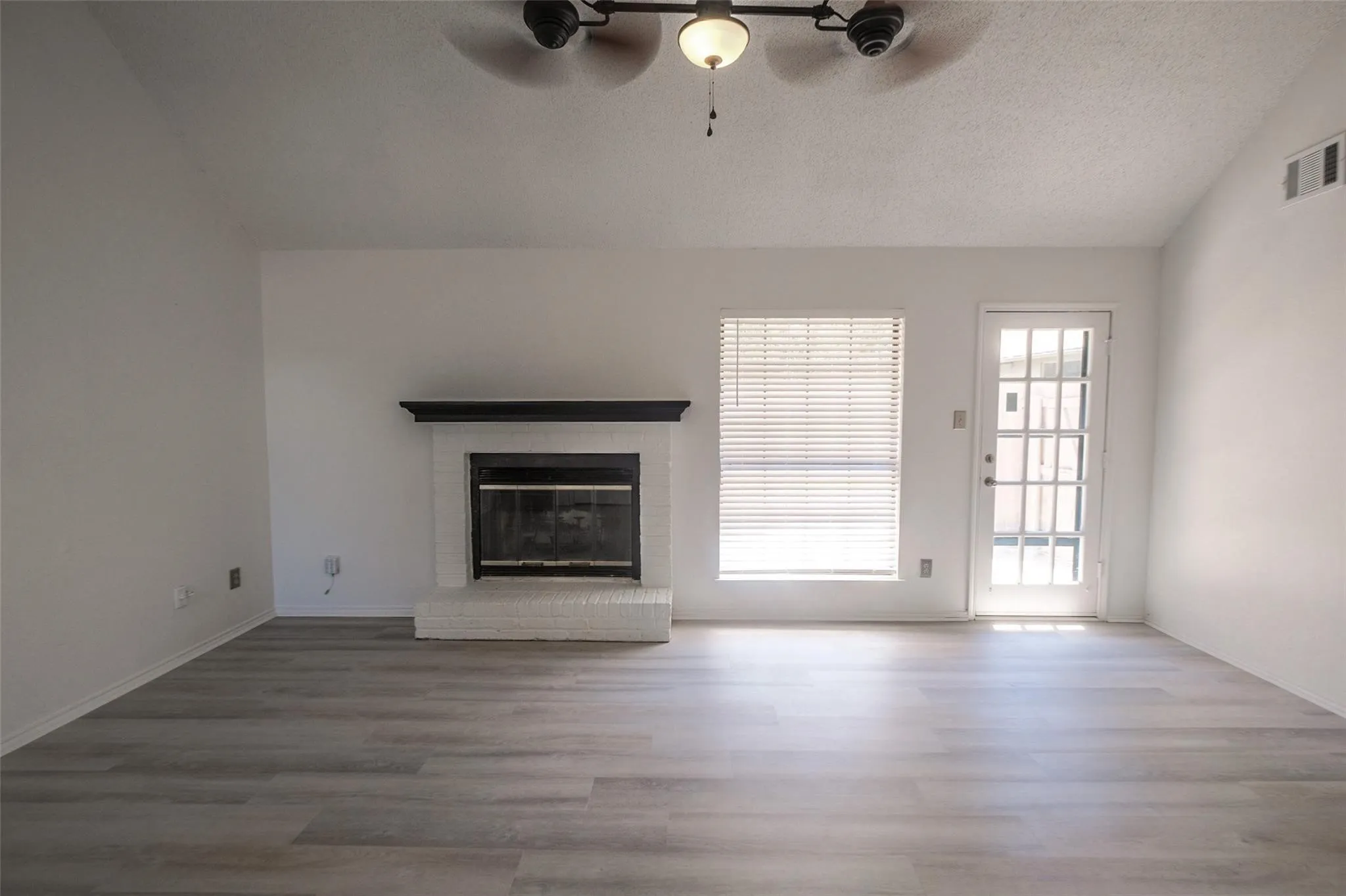 Wood-finish flooring, white brick fireplace with dark mantel, window with blinds, ceiling fan, and exterior door with glass panels