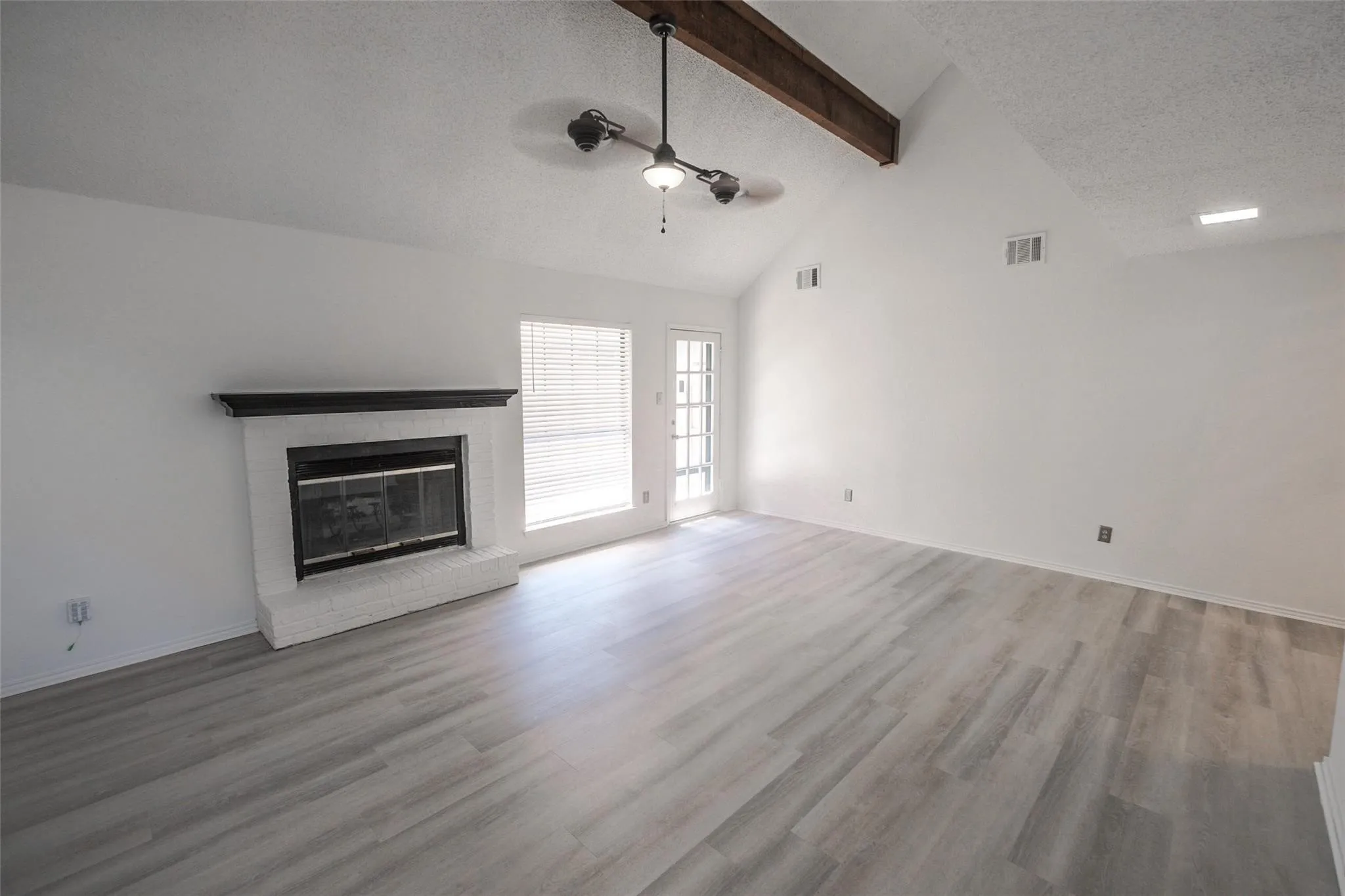 Vaulted ceiling with exposed wood beam, modern ceiling fan, wood-finish flooring, and a white brick fireplace with a black mantel