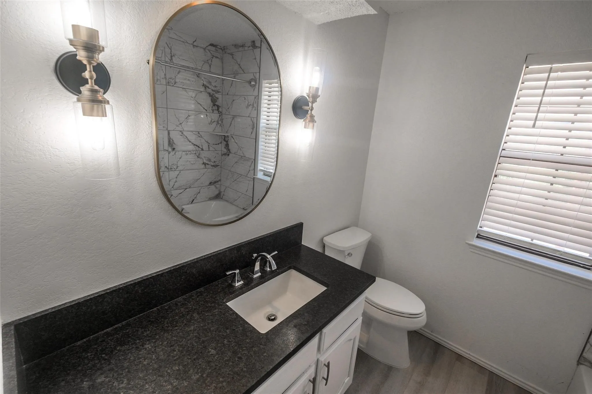 Vanity with a dark countertop and integrated sink, oval mirror, and wall-mounted sconces