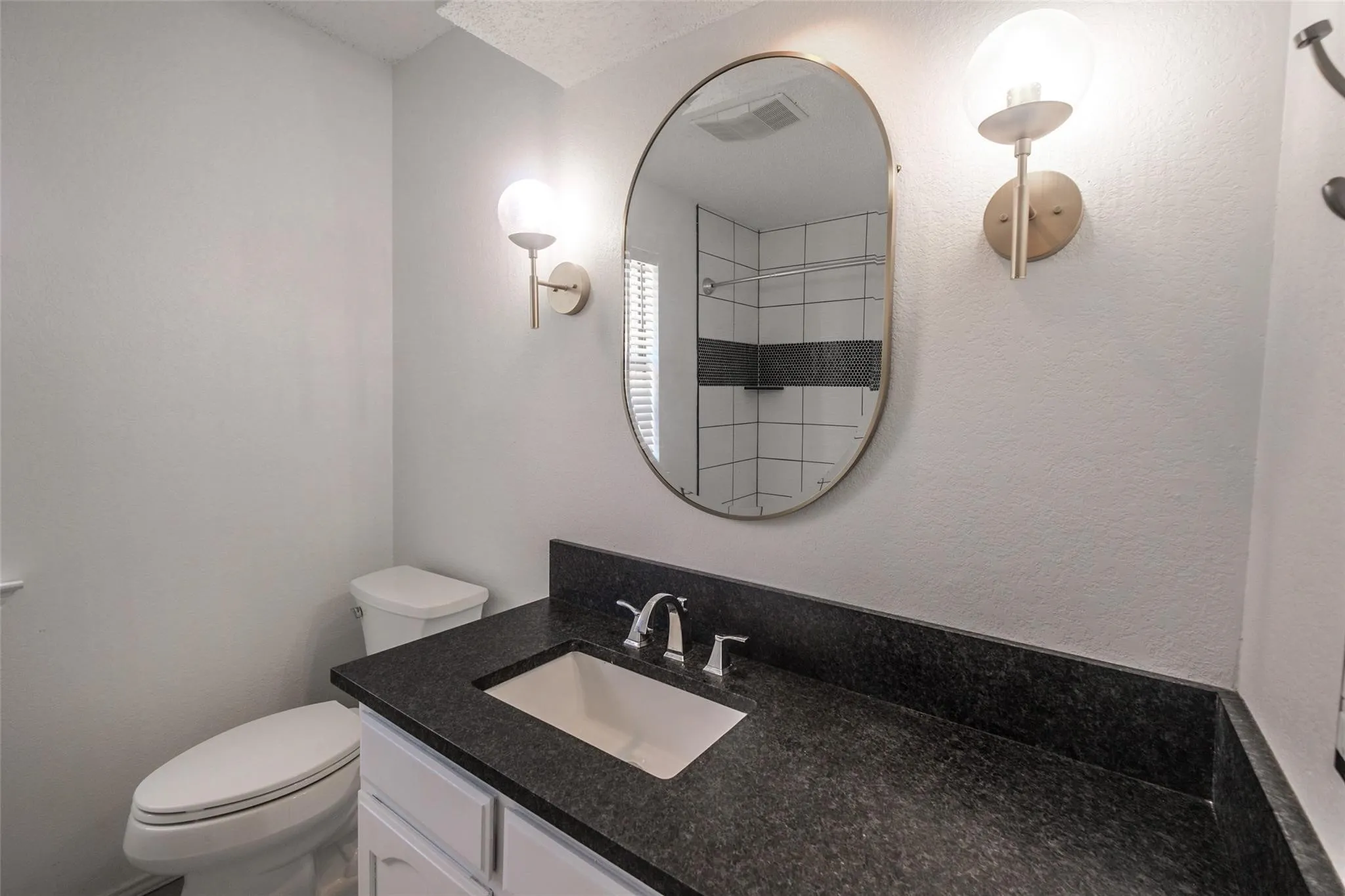 Dark countertop with an integrated rectangular sink, white vanity cabinetry, brushed gold sconces, and an oval mirror