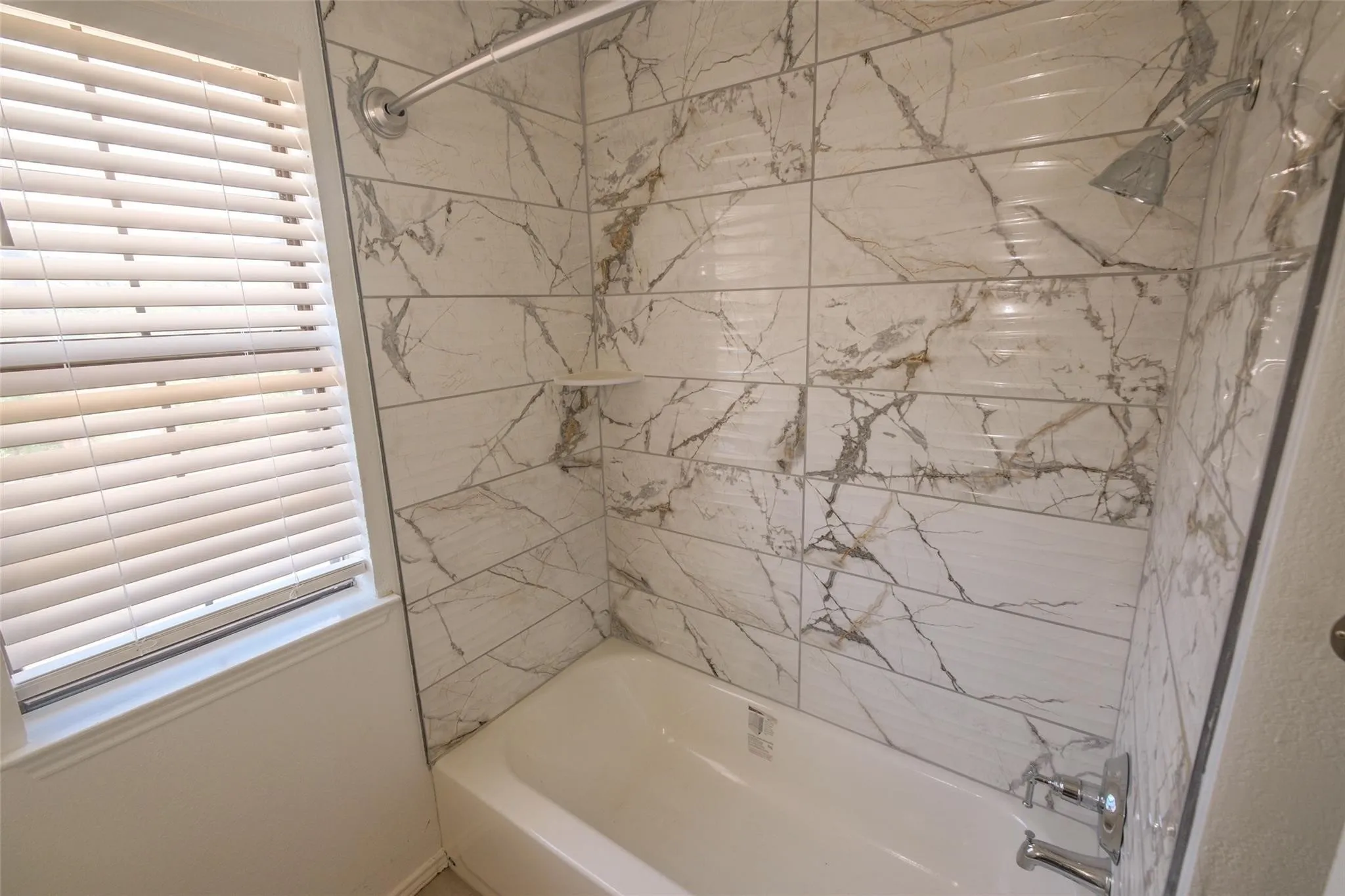 Bathtub with chrome fixtures, tiled shower surround with integrated shelf, and window with blinds
