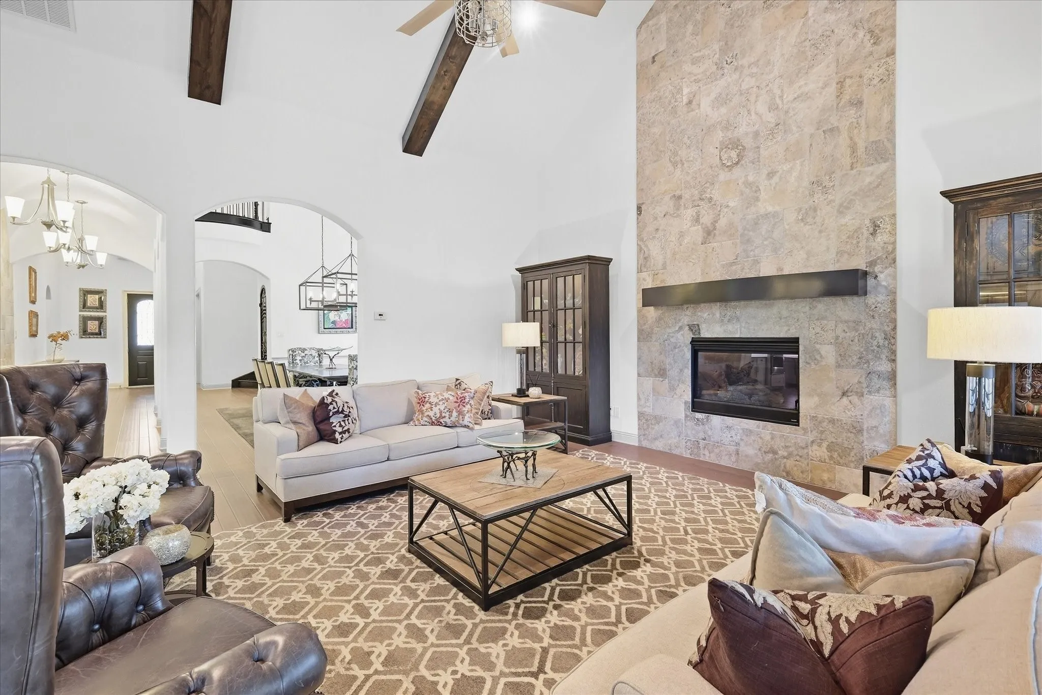 Soaring vaulted ceilings with dark wood beams, expansive stone fireplace with a dark wood mantel, integrated electric fireplace, arched doorways, and wood flooring
