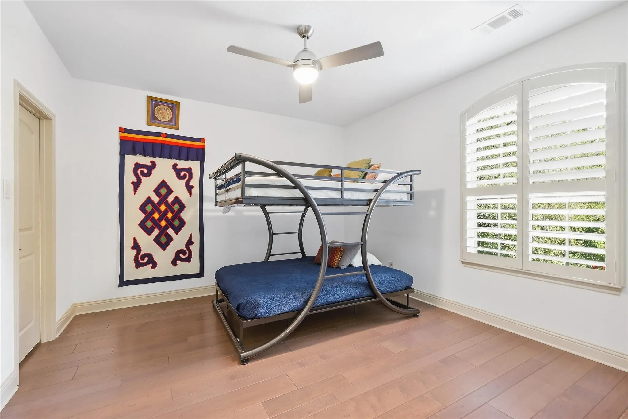 Wood flooring, white walls, arched window with plantation shutters, ceiling fan with integrated lighting, and a white interior door
