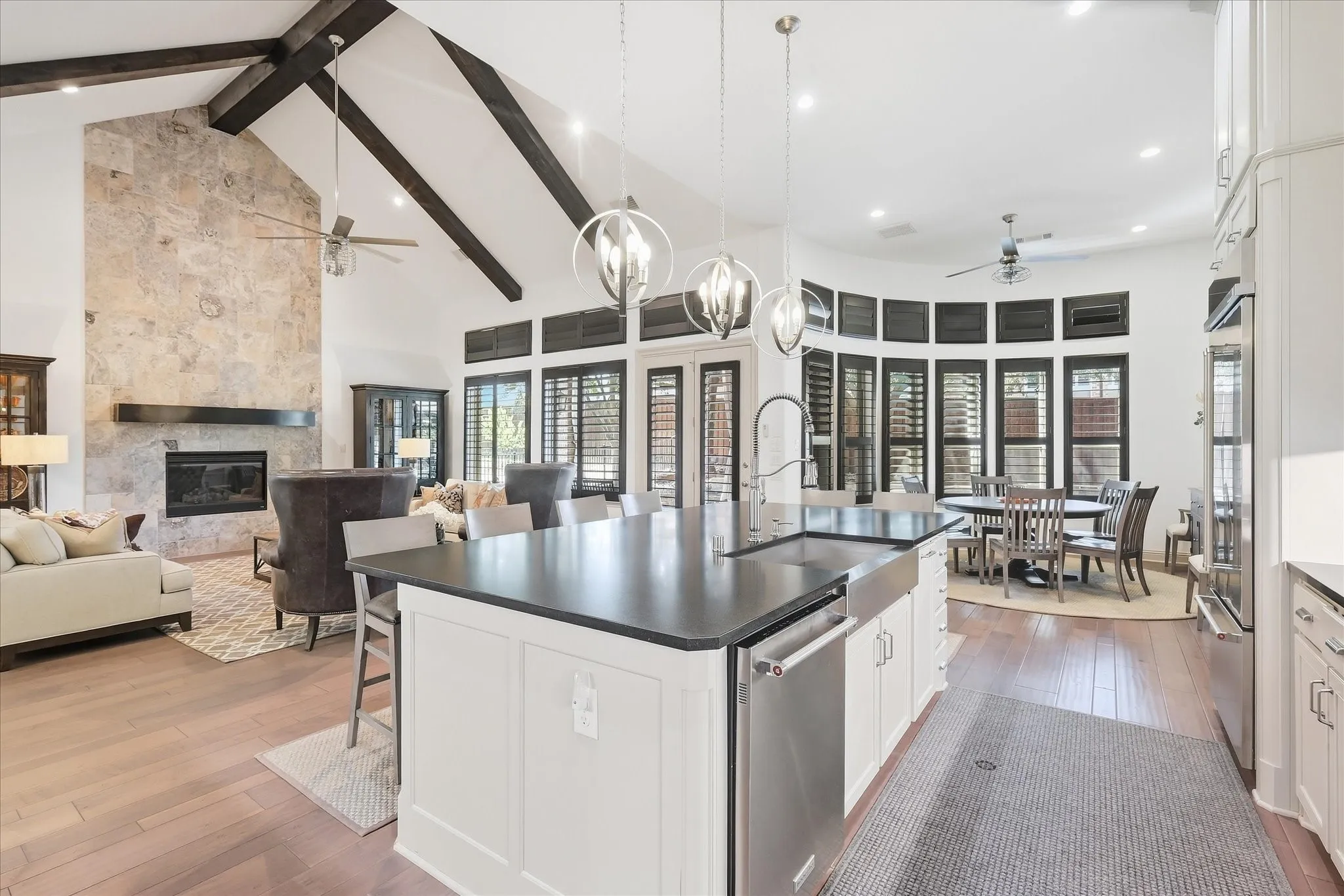 Vaulted ceiling with exposed beams, stone fireplace, wood-finish flooring, white kitchen cabinetry with dark countertops, and a bay window dining area