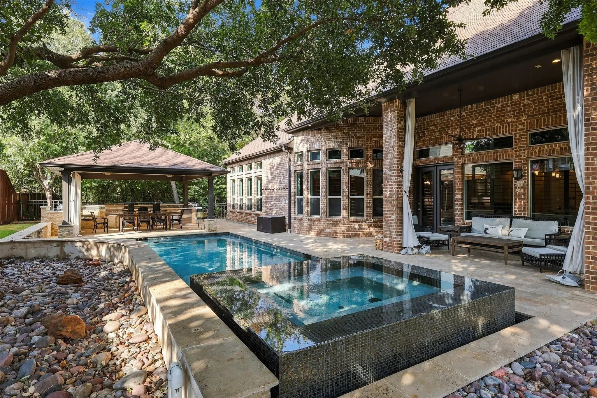 Outdoor living area featuring a rectangular pool with an integrated spa, expansive brick exterior, covered patio, and a separate covered outdoor kitchen
