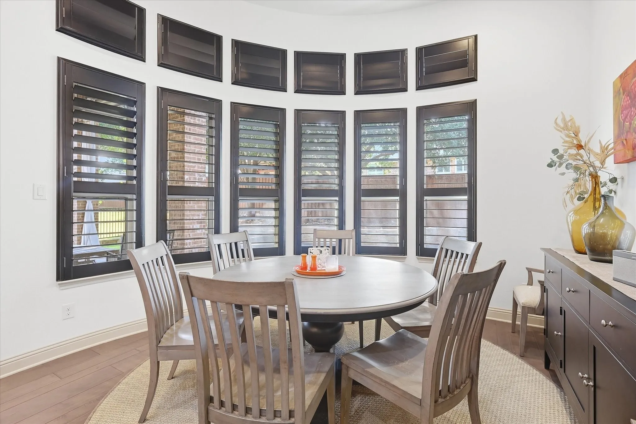 Curved bay window with dark plantation shutters, light wood-finish flooring, and a built-in dark wood credenza with light countertop
