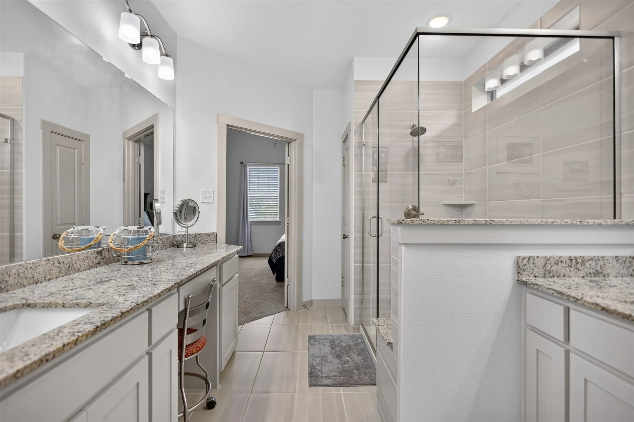 Dual vanity with granite countertops, glass-enclosed shower, tiled flooring, and modern light fixtures