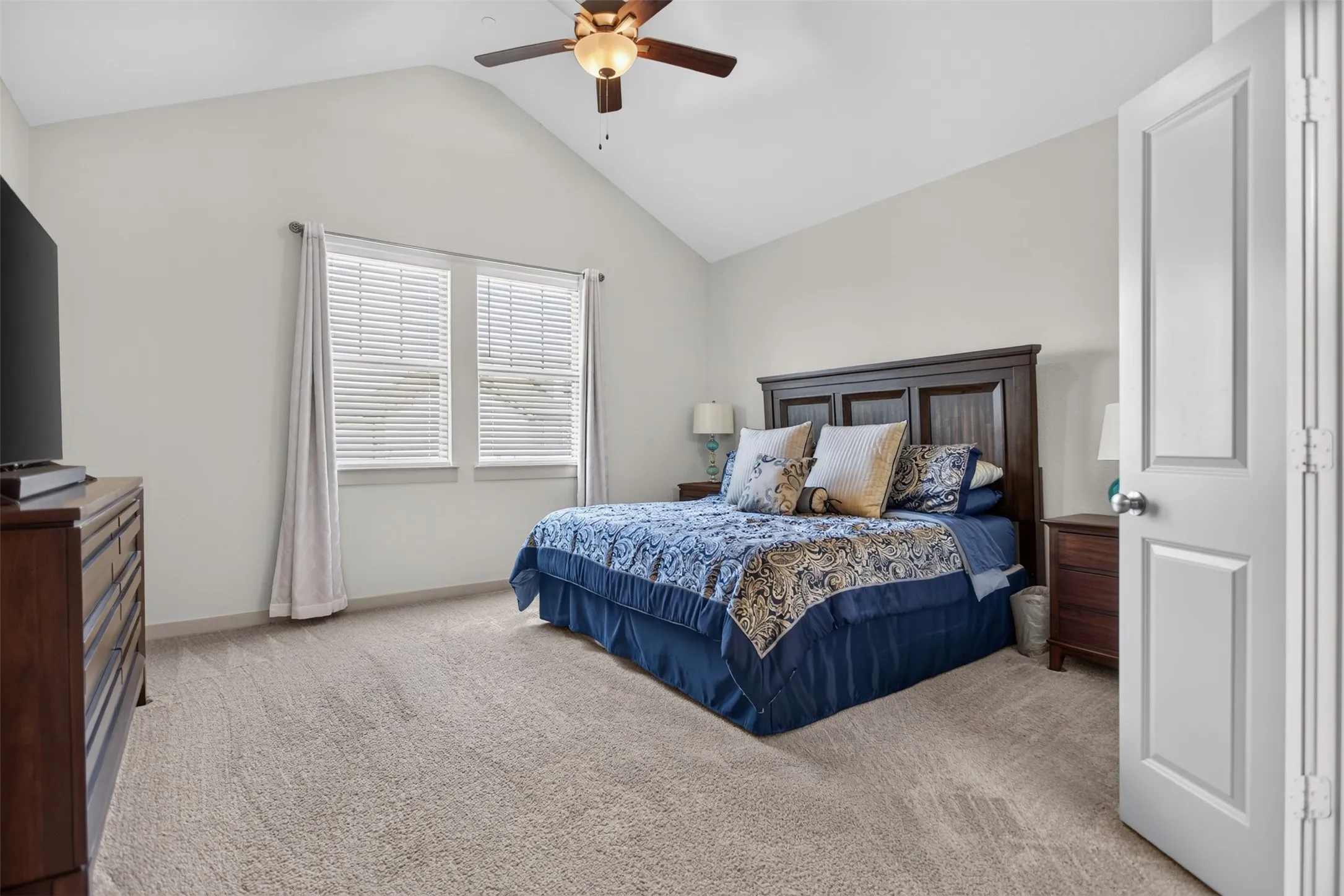 Vaulted ceiling with a ceiling fan, two windows with blinds, carpeted flooring, and a paneled door