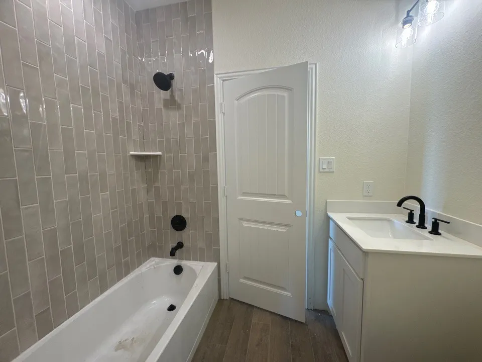 Bathtub with subway tile surround, matte black shower fixtures, wood-finish flooring, white vanity with integrated sink, and matte black faucet