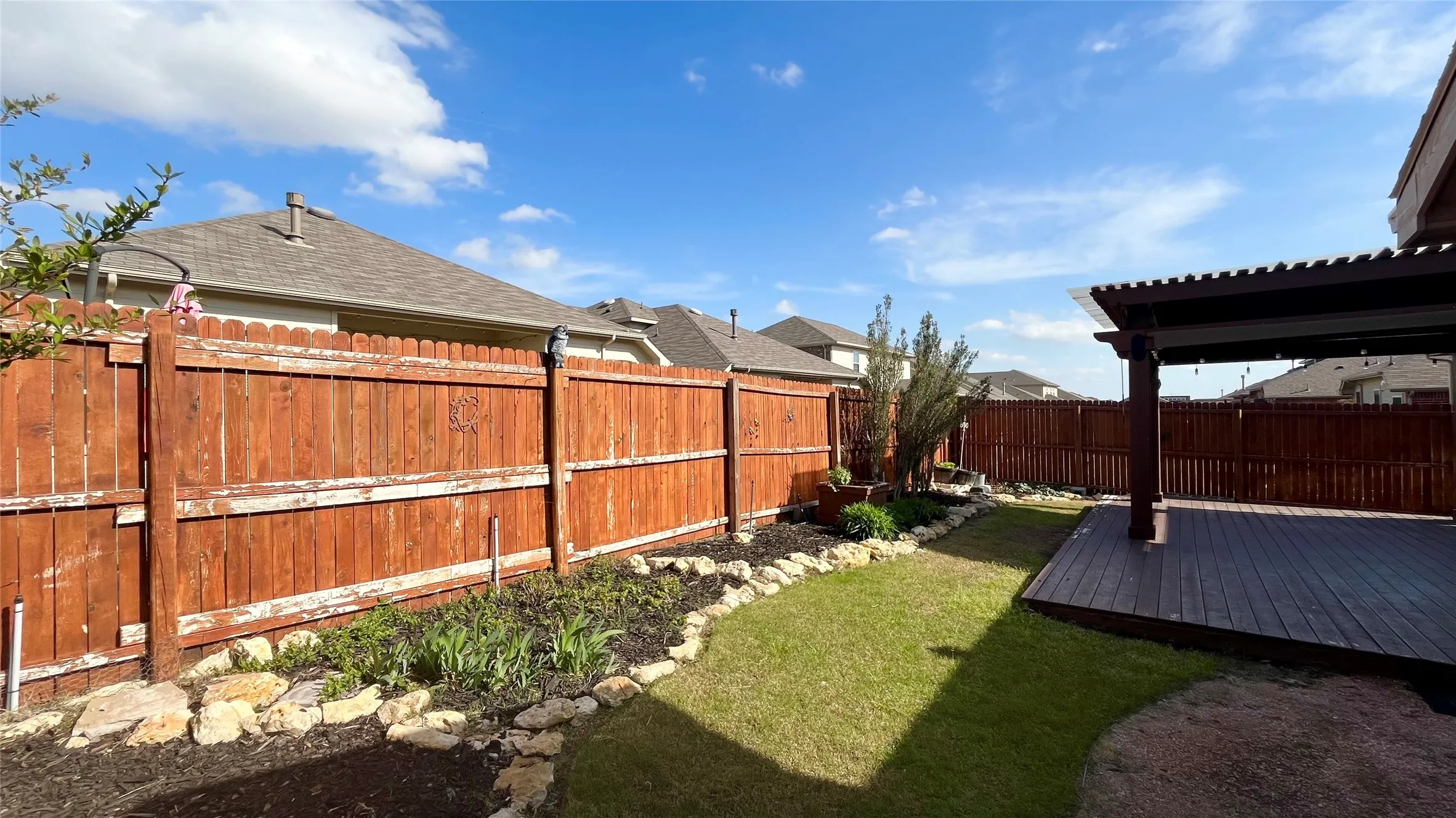 Expansive backyard featuring a manicured lawn, a large wood-finish deck, a covered pergola, a rock-lined garden bed, and privacy fencing