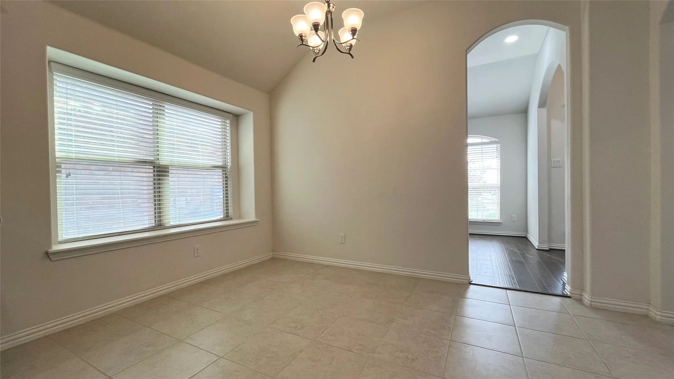 Vaulted ceiling with an attached chandelier, large window with blinds, tile flooring, and arched entryway