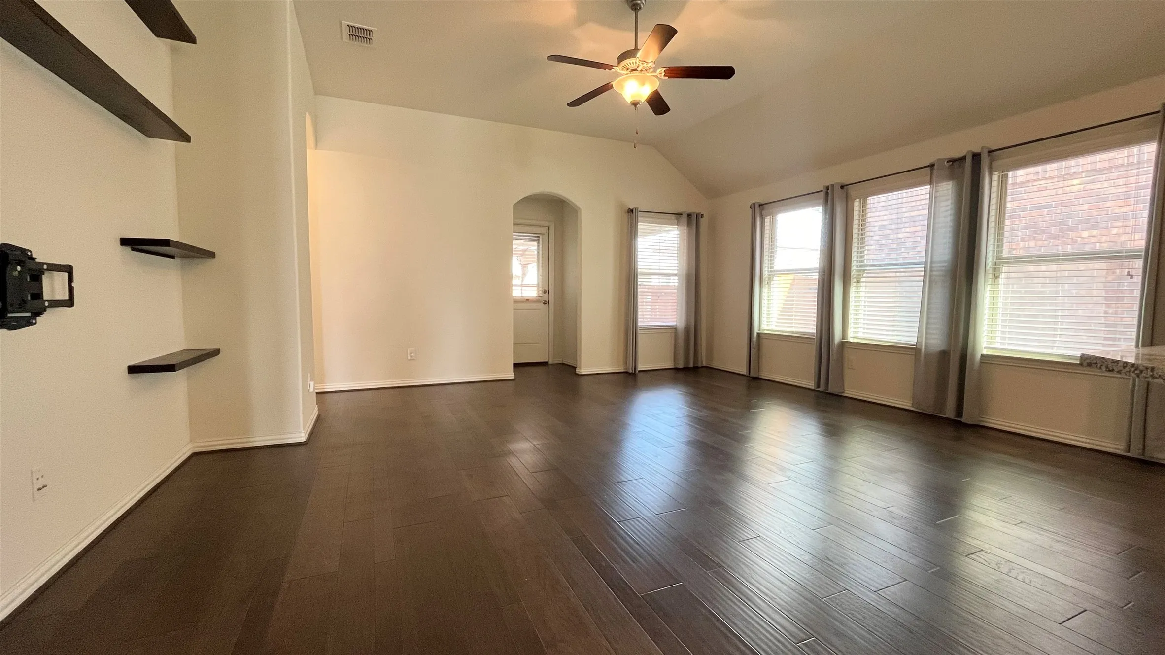 Vaulted ceiling, wood-finish flooring, ceiling fan with integrated lighting, multiple windows with blinds, and built-in wall shelving