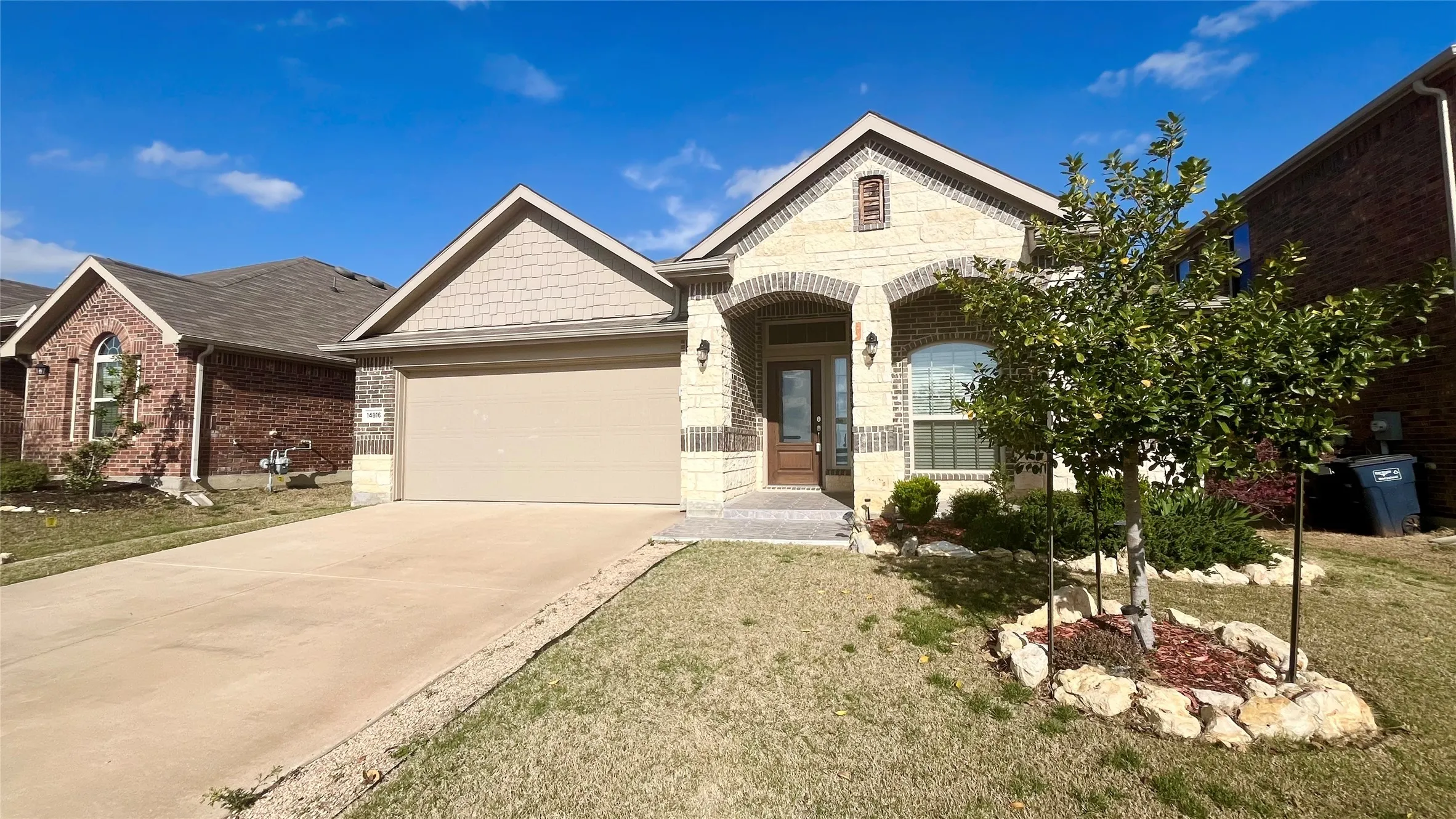 Brick and stone facade, arched entry, mature landscaping, paved driveway, and an attached two-car garage