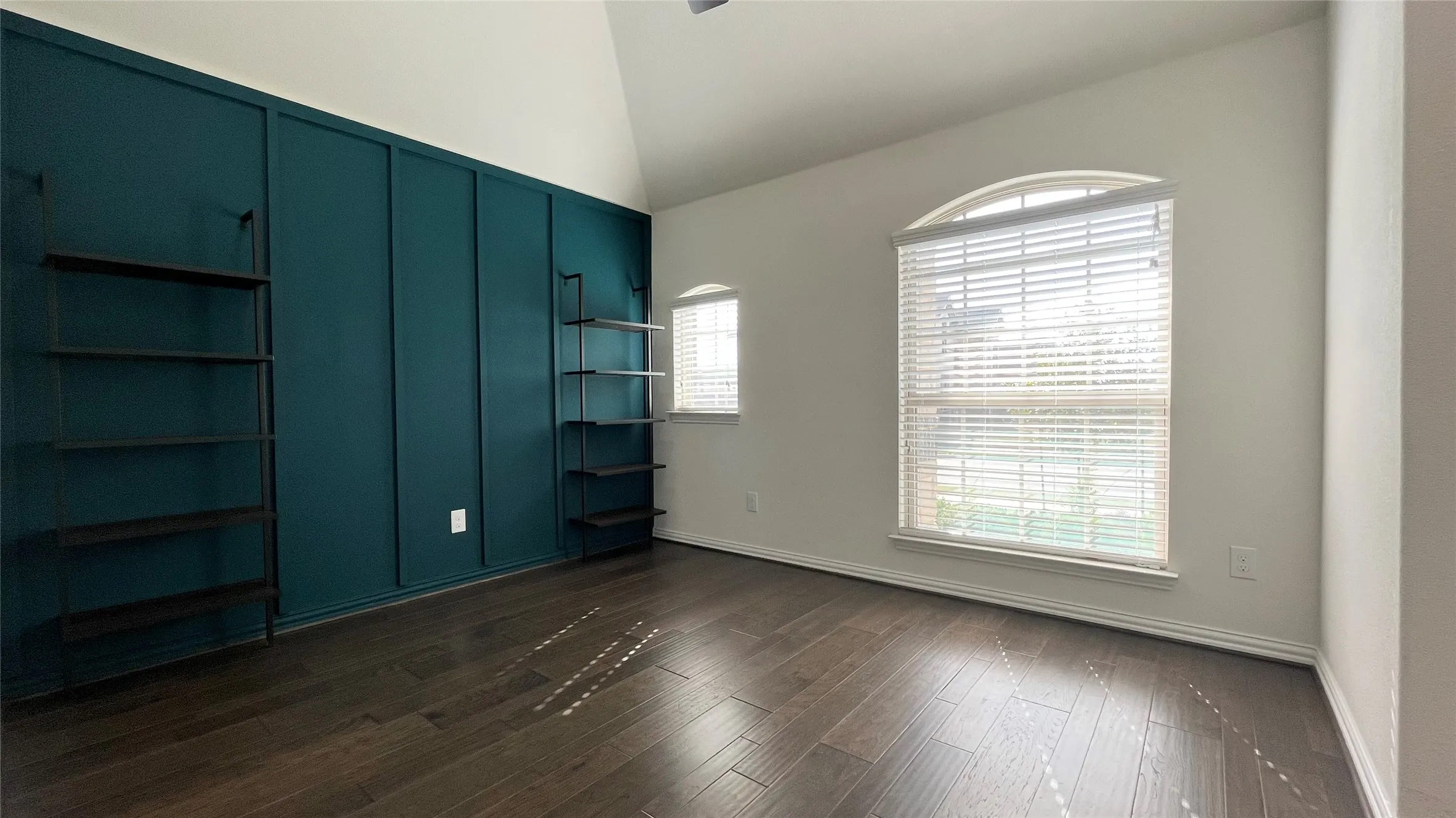 Vaulted ceiling, wood-finish flooring, accent wall with paneling, built-in shelving, and an arched window