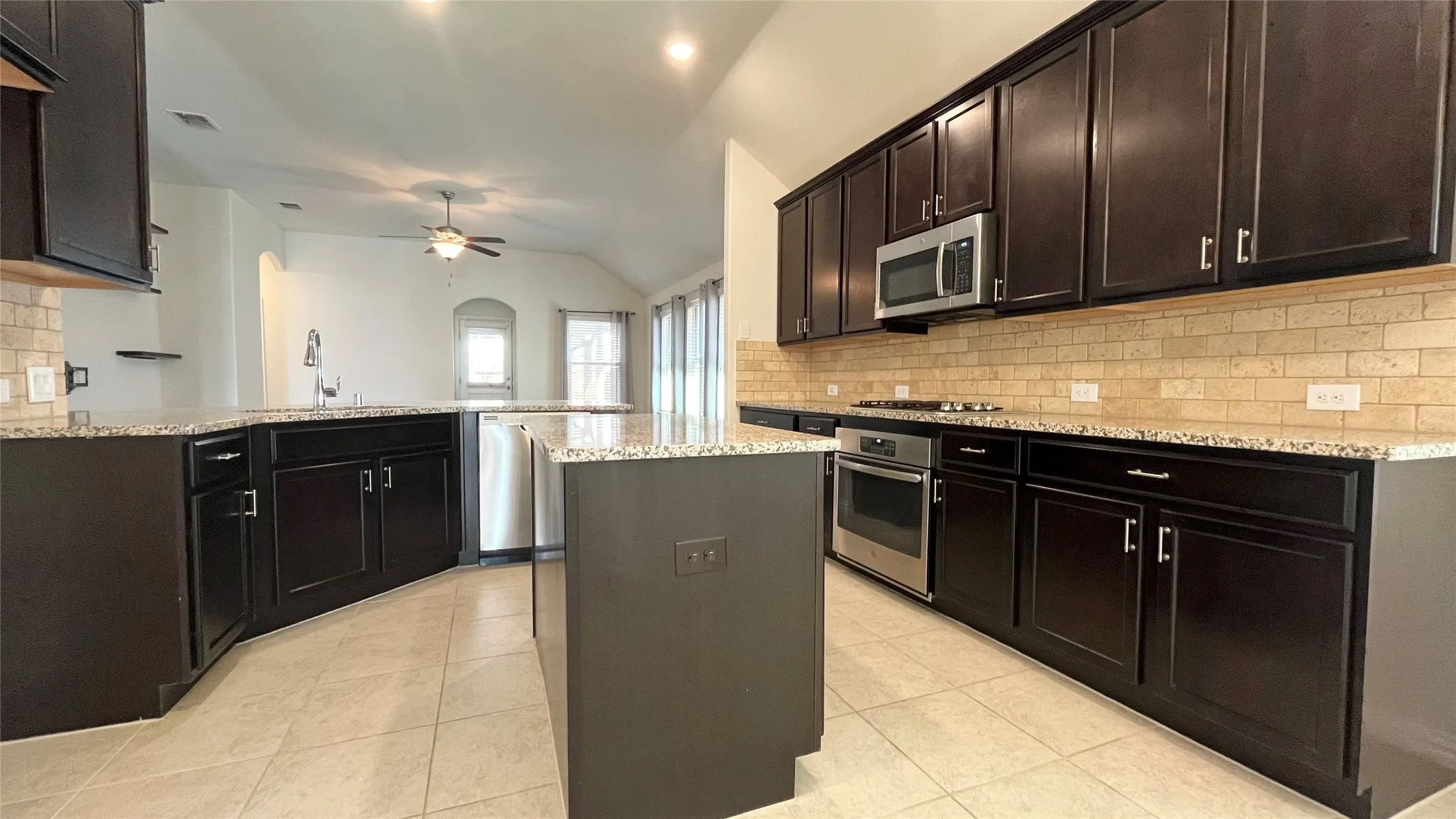 Spacious kitchen featuring dark wood cabinetry, light-toned tiled backsplash, stainless steel appliances, and a central island with a light-toned countertop