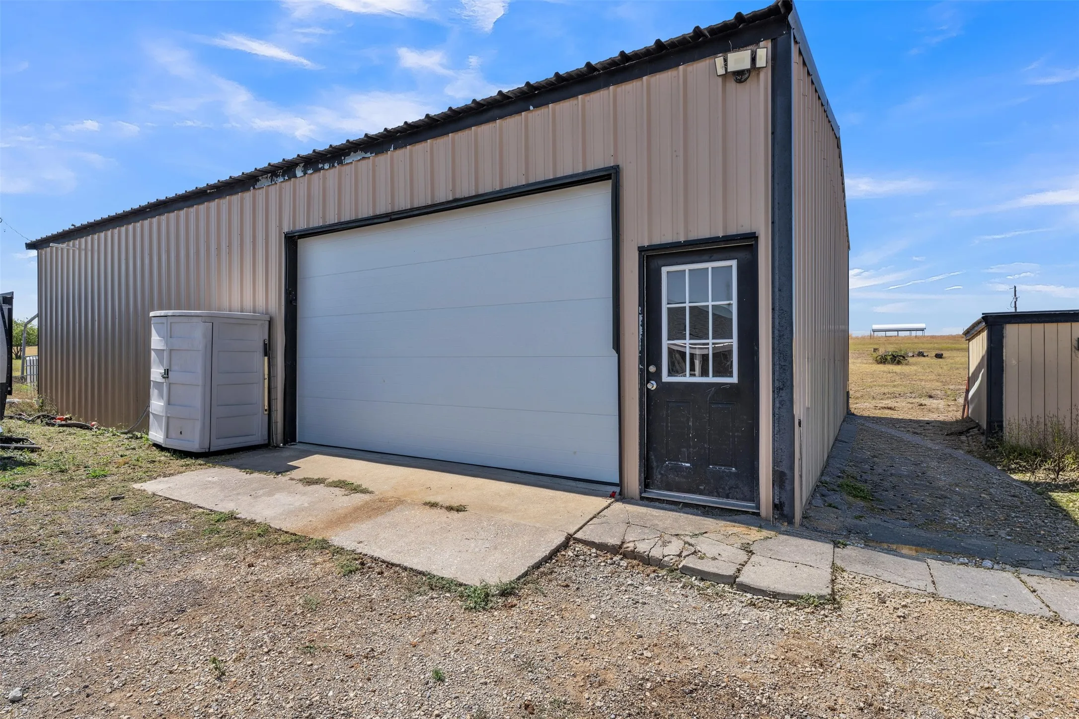 Corrugated metal building featuring a large white roll-up door, a black paneled entry door with window panes, and an exterior light fixture