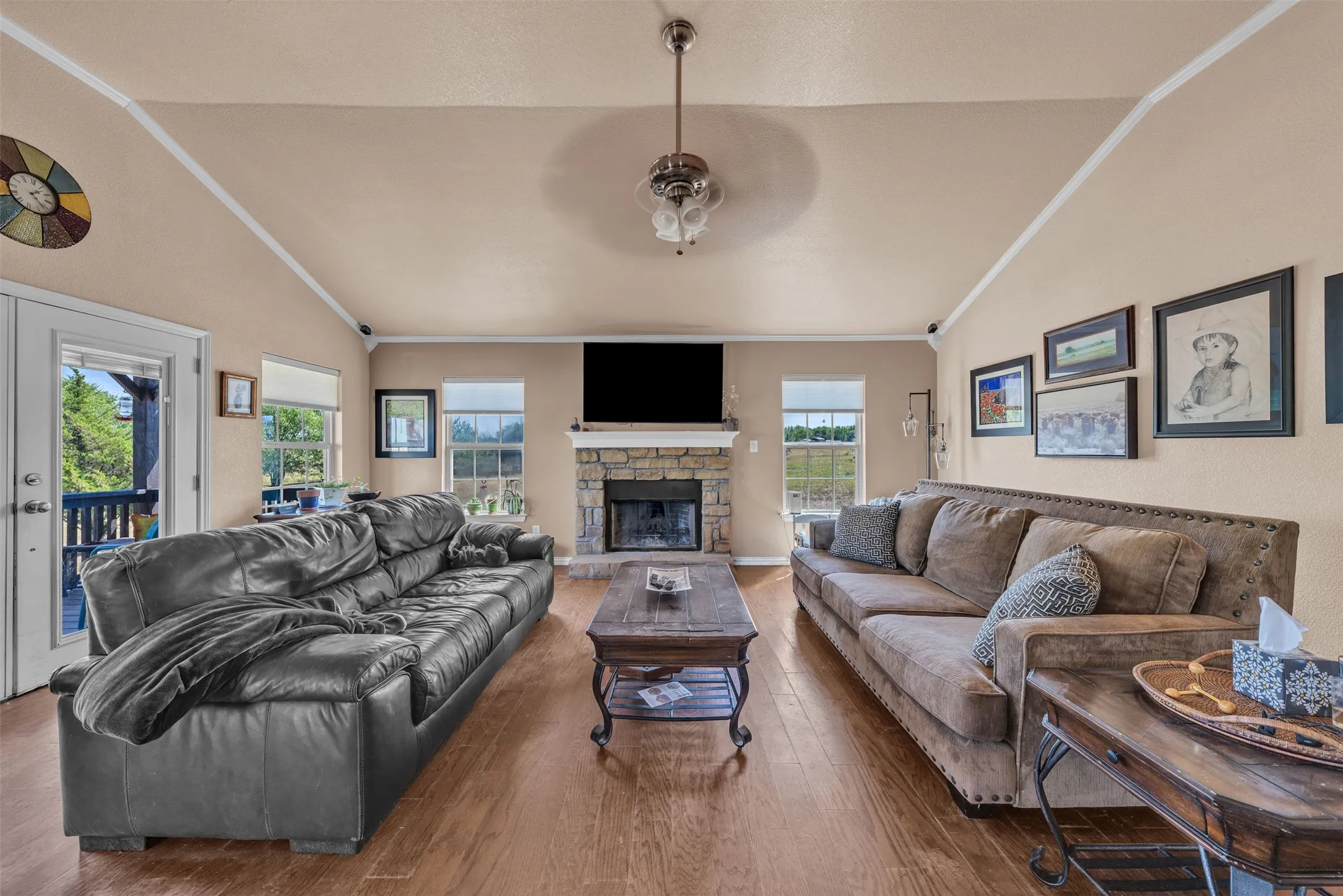 Vaulted ceiling, wood-finish flooring, stone-surround fireplace, ceiling fan, and multiple windows