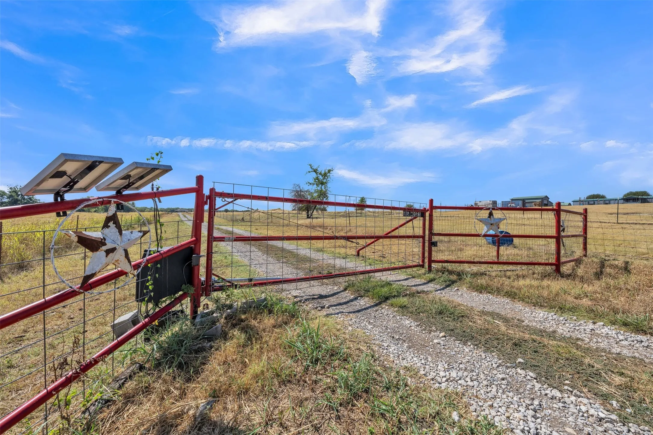 Red metal gate with wire fencing, gravel driveway, and solar panels