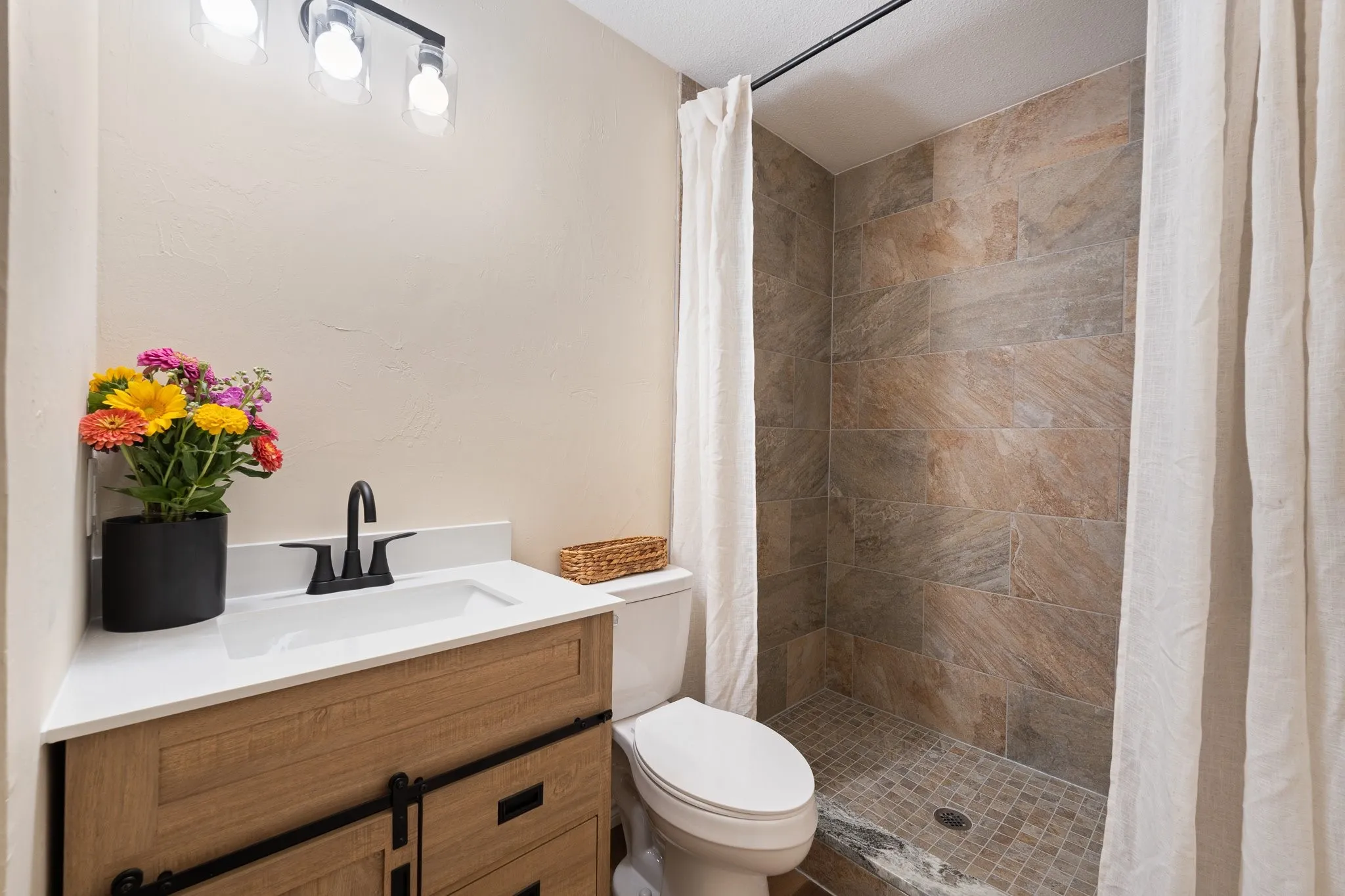 Walk-in shower with textured tile surround, wood-finish vanity with sliding barn door style cabinet fronts, white countertop with integrated sink, and black fixtures