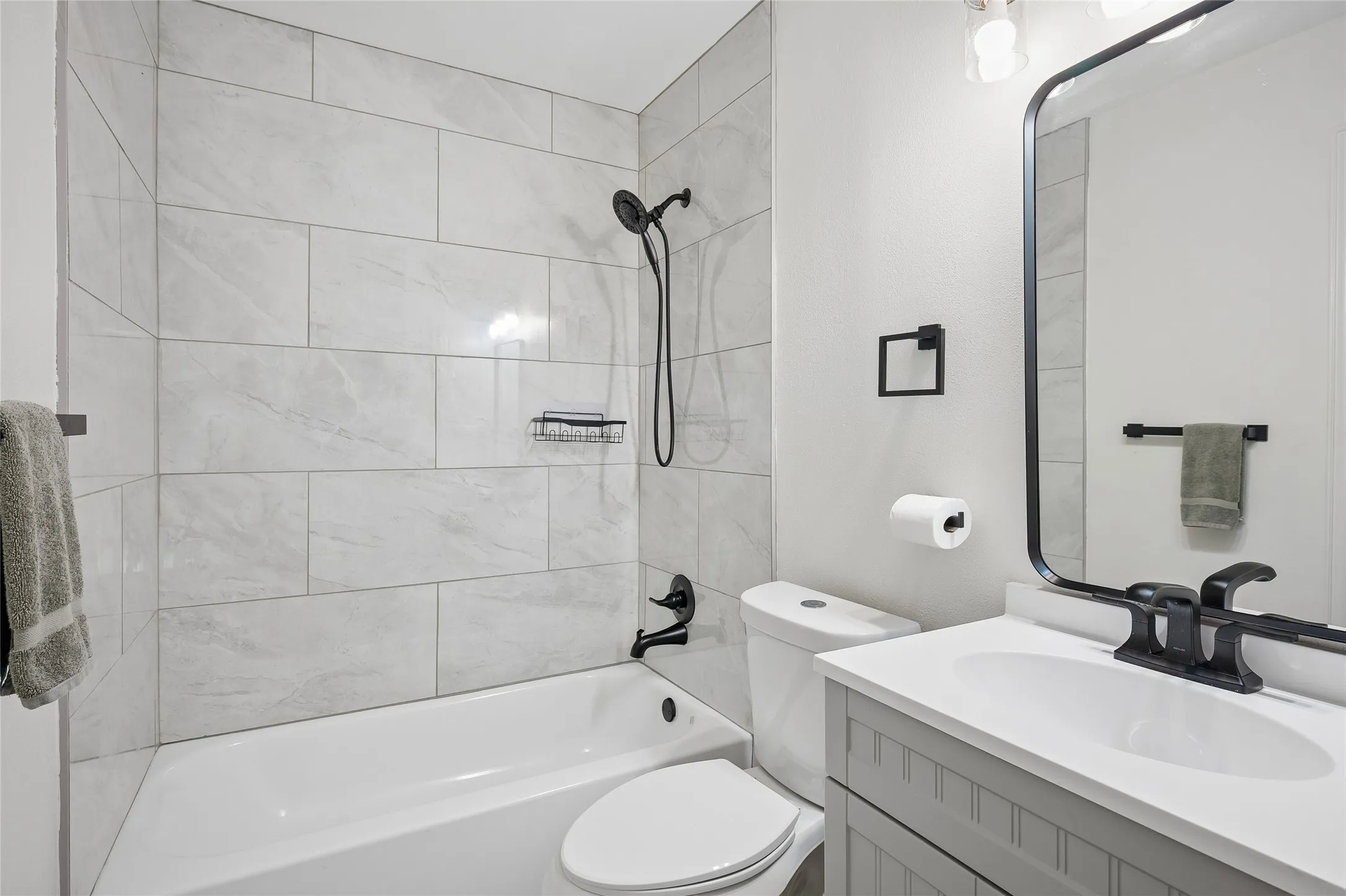 Bathroom featuring a white bathtub, large format tile shower surround, matte black fixtures, a single vanity with a white countertop, and a framed mirror