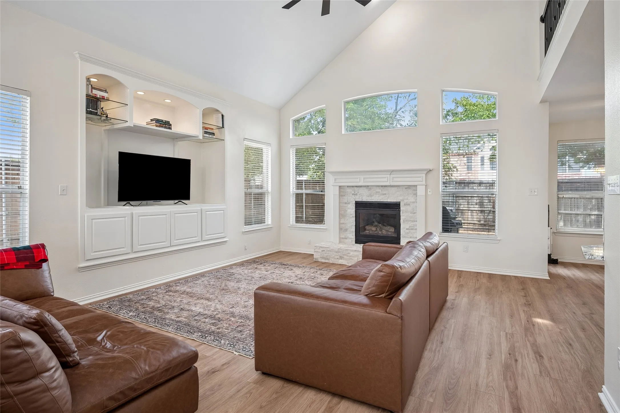 Vaulted ceiling with a ceiling fan, built-in entertainment center with shelving, fireplace with a tiled surround and mantel, abundant natural light from multiple windows, and wood-finish flooring